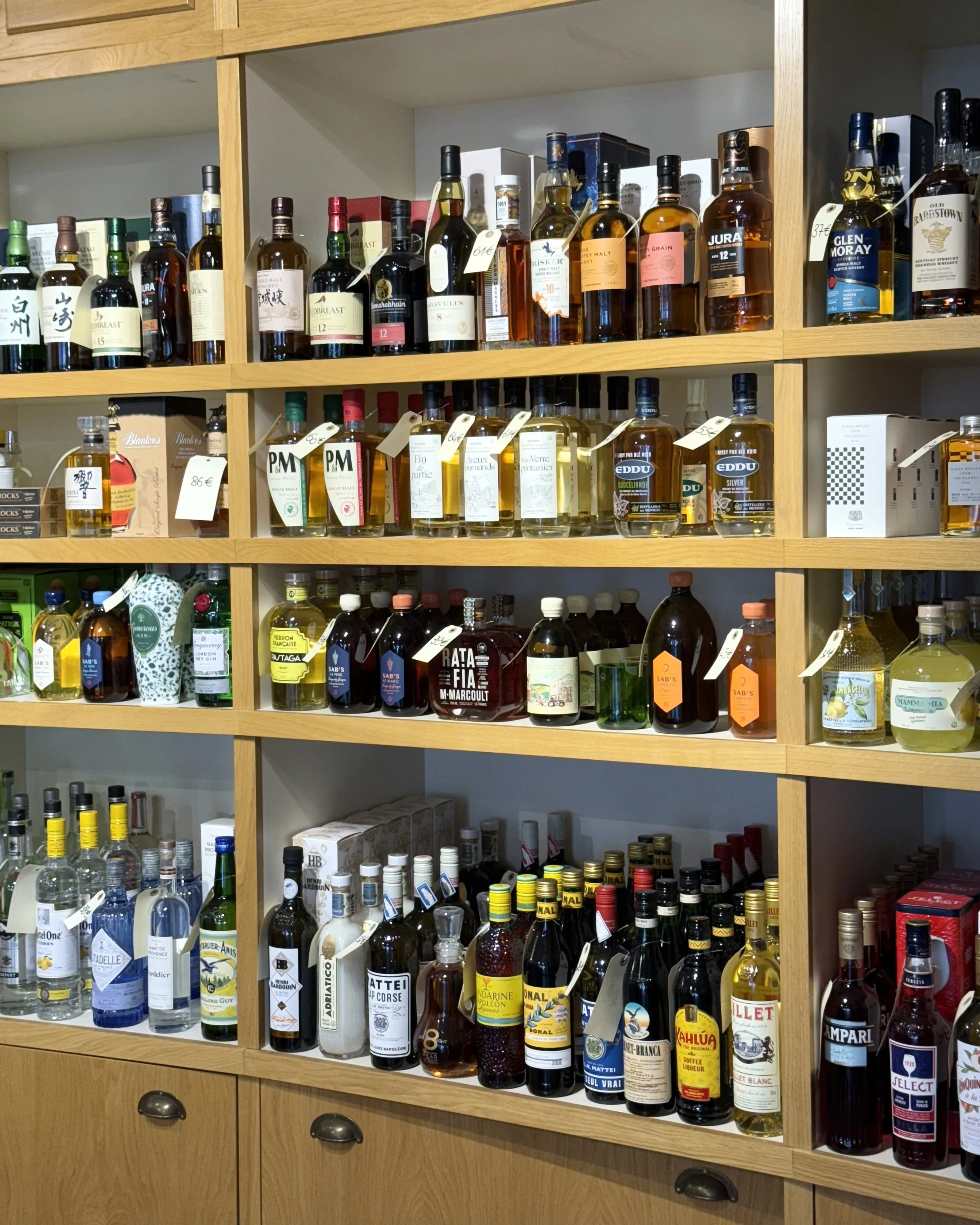 Étagères en bois remplies de diverses bouteilles d'alcool, notamment des spiritueux, des liqueurs et des whiskys, avec des étiquettes manuscrites ou imprimées.