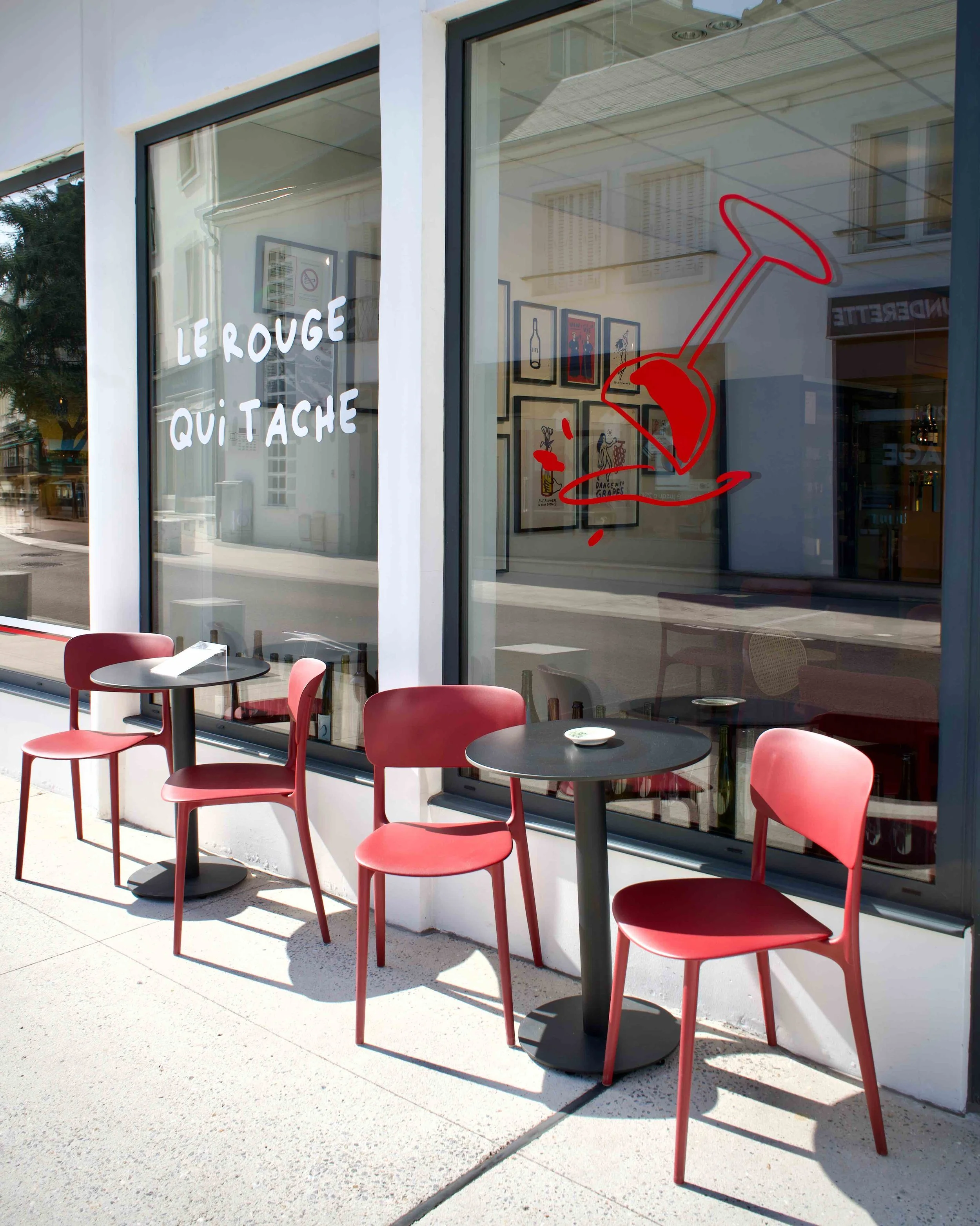 Deux tables rondes avec des chaises rouges à l'extérieur d'un café en terrasse à Vernon, vue à travers une grande vitre. Sur la vitre, une illustration d'une bouteille de vin, un verre,  'Le rouge qui tache' écrit en blanc.