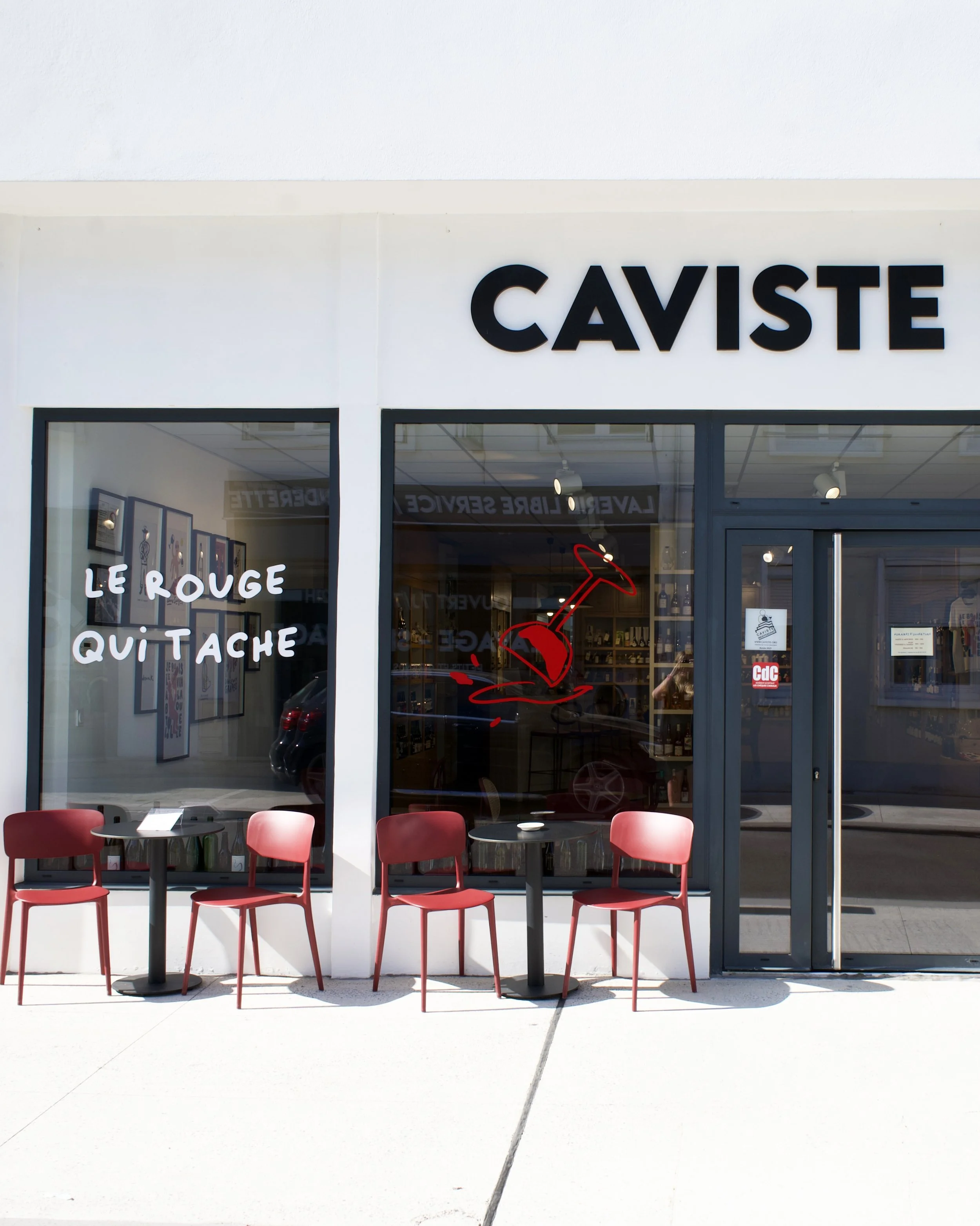 Facade de magasin avec le nom "CAVISTE", fenêtres avec message "LE ROUGE QUI TACHE", et devant une terrasse avec trois chaises rouges et deux tables dans la ville de Vernon à côté de Giverny dans l'Eure en Normandie