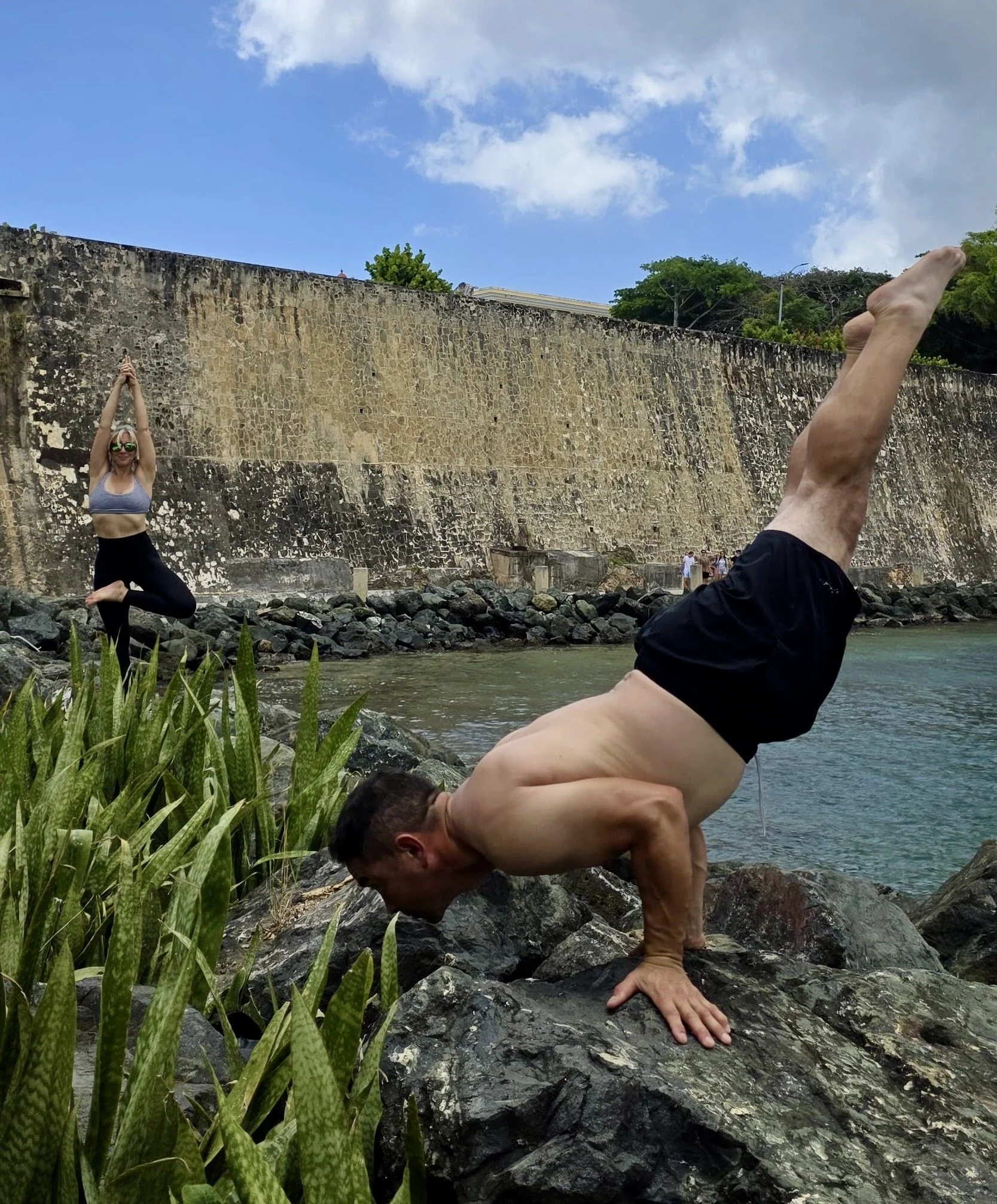 A man doing a handstand on rocks near a water body with a woman practicing yoga or meditation in a tree pose in the background, against a stone wall and blue sky with white clouds.
