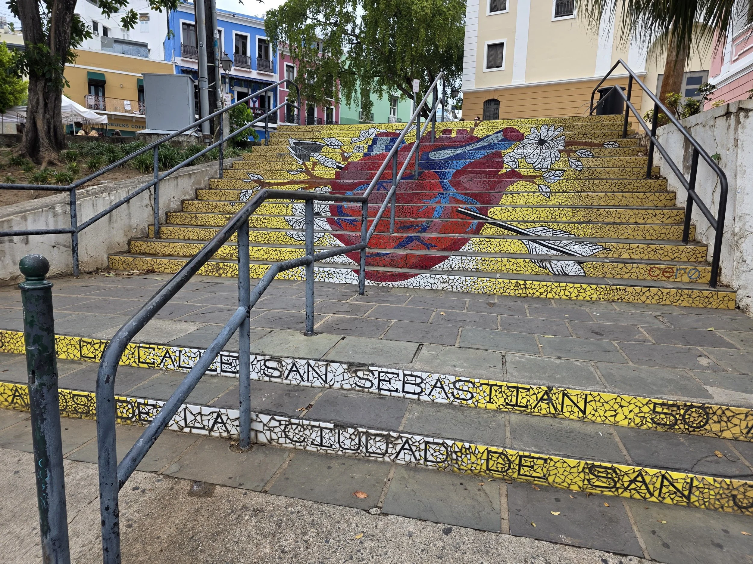 Colorful painted stairs depicting a large red heart with white petals, surrounded by green and yellow mosaic tiles, in an urban setting with buildings in pastel colors in the background.