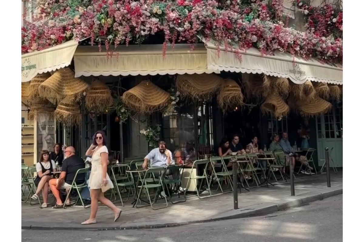 People sitting outside a café with pink and white flowers on the building's awning, some using phones, one woman in a white dress walking by.