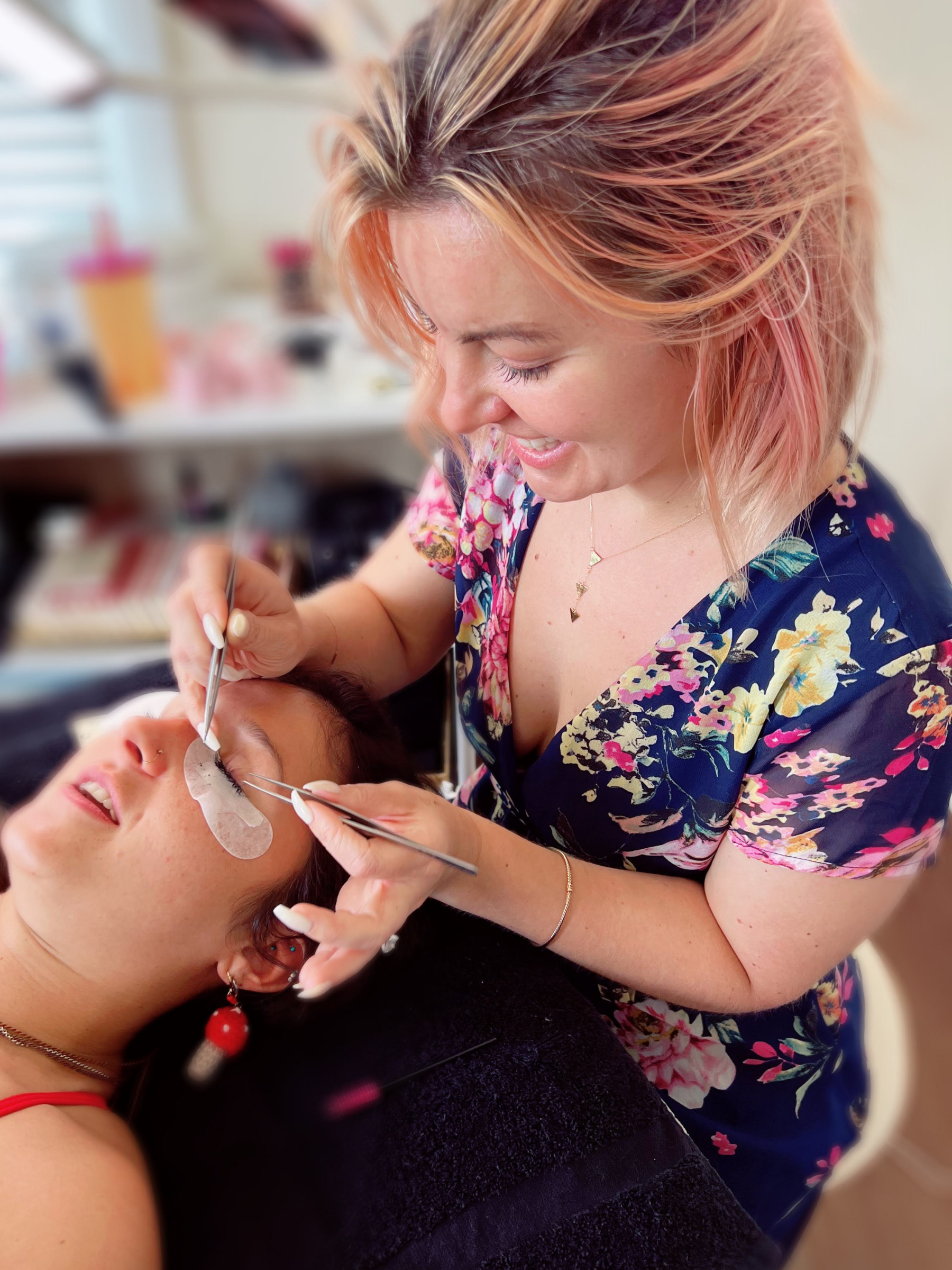 A woman with pinkish-orange hair getting a facial treatment from a professional in a floral dress, in a bright room with various beauty supplies in the background.