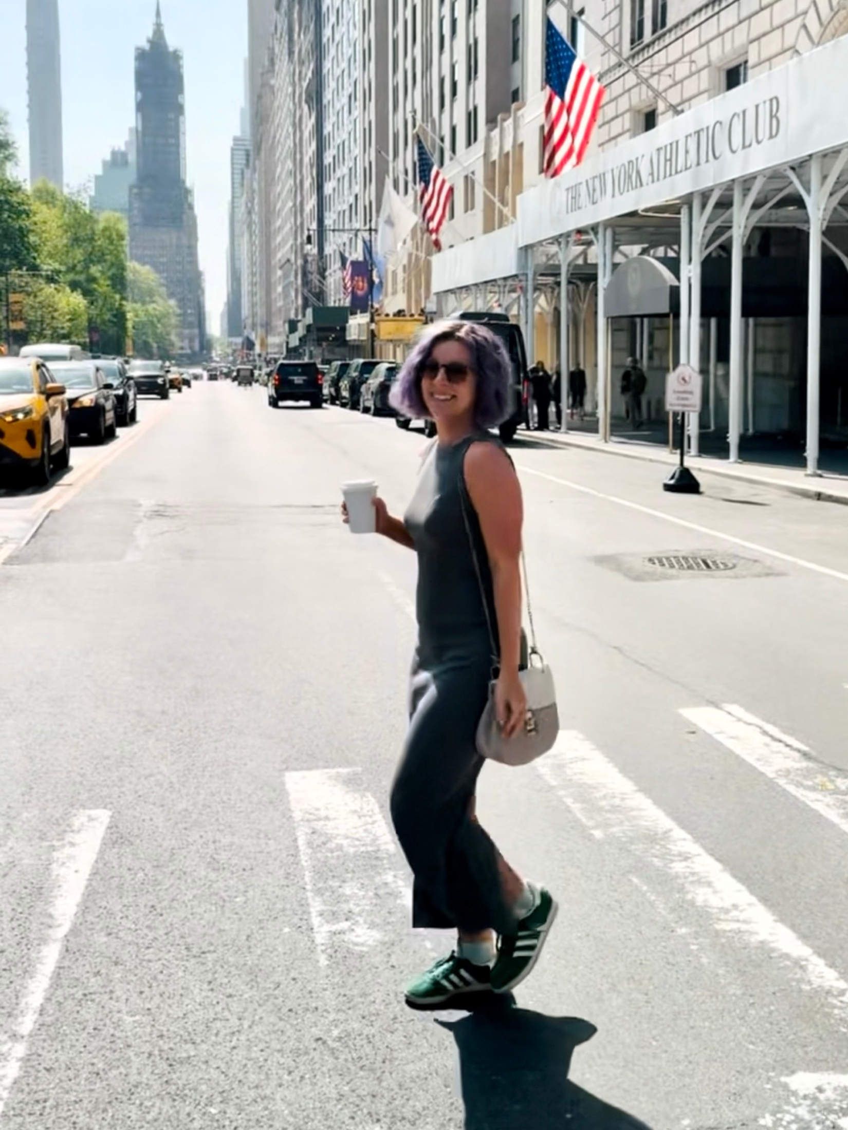 A smiling woman with purple hair and sunglasses crossing a city street with tall buildings, parked cars, and flags, including American flags, on a building with a sign for The New York Athletic Club.