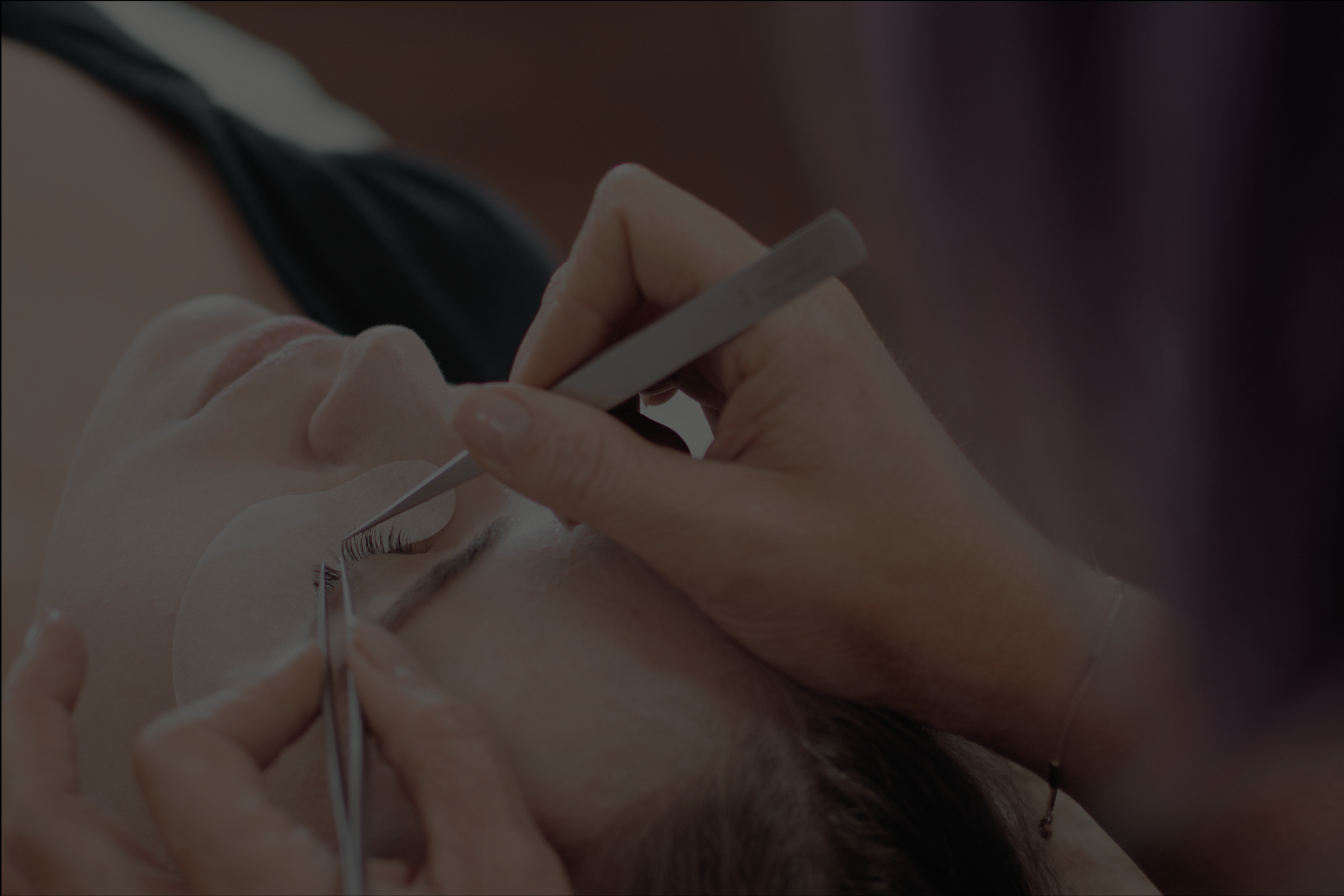 A close-up of a person receiving eyelash extension treatment, with an aesthetician applying eyelash extensions using tweezers while the client has eye pads under their eyes.