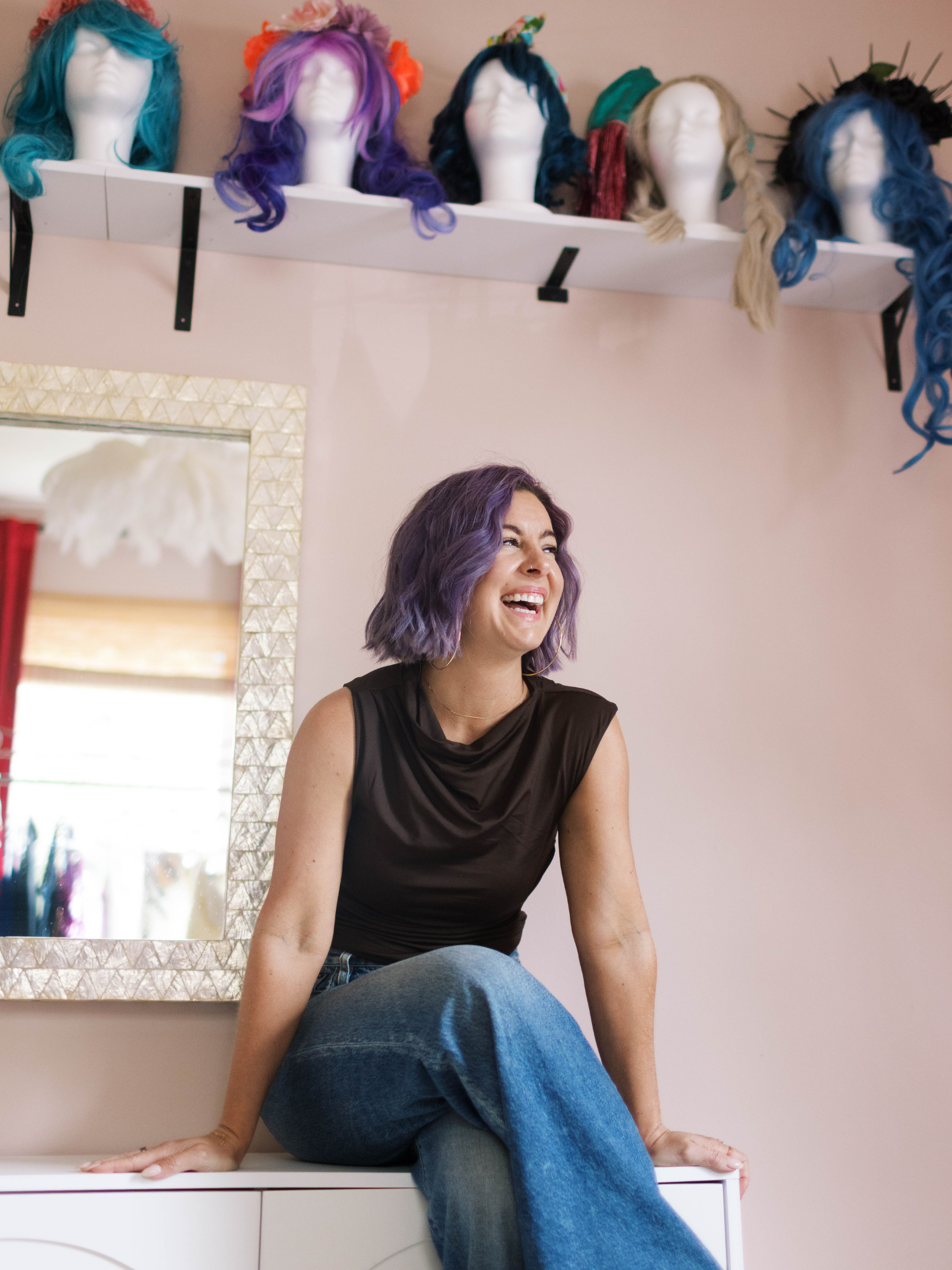 A woman with purple hair sitting on a white surface, smiling, with wigs on mannequin heads mounted on a shelf above her.