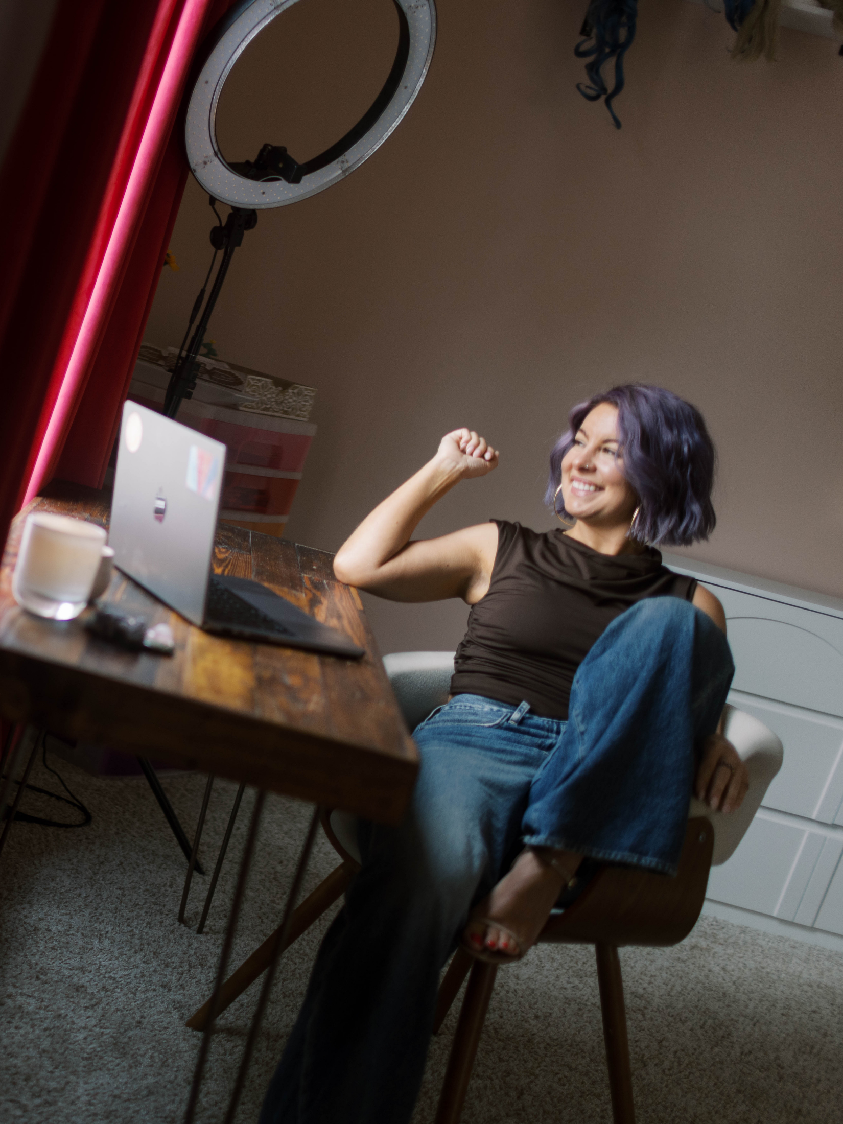 A woman with purple hair smiling and flexing her arm, sitting at a wooden table in a room with a ring light overhead, a laptop, and a glass of water.