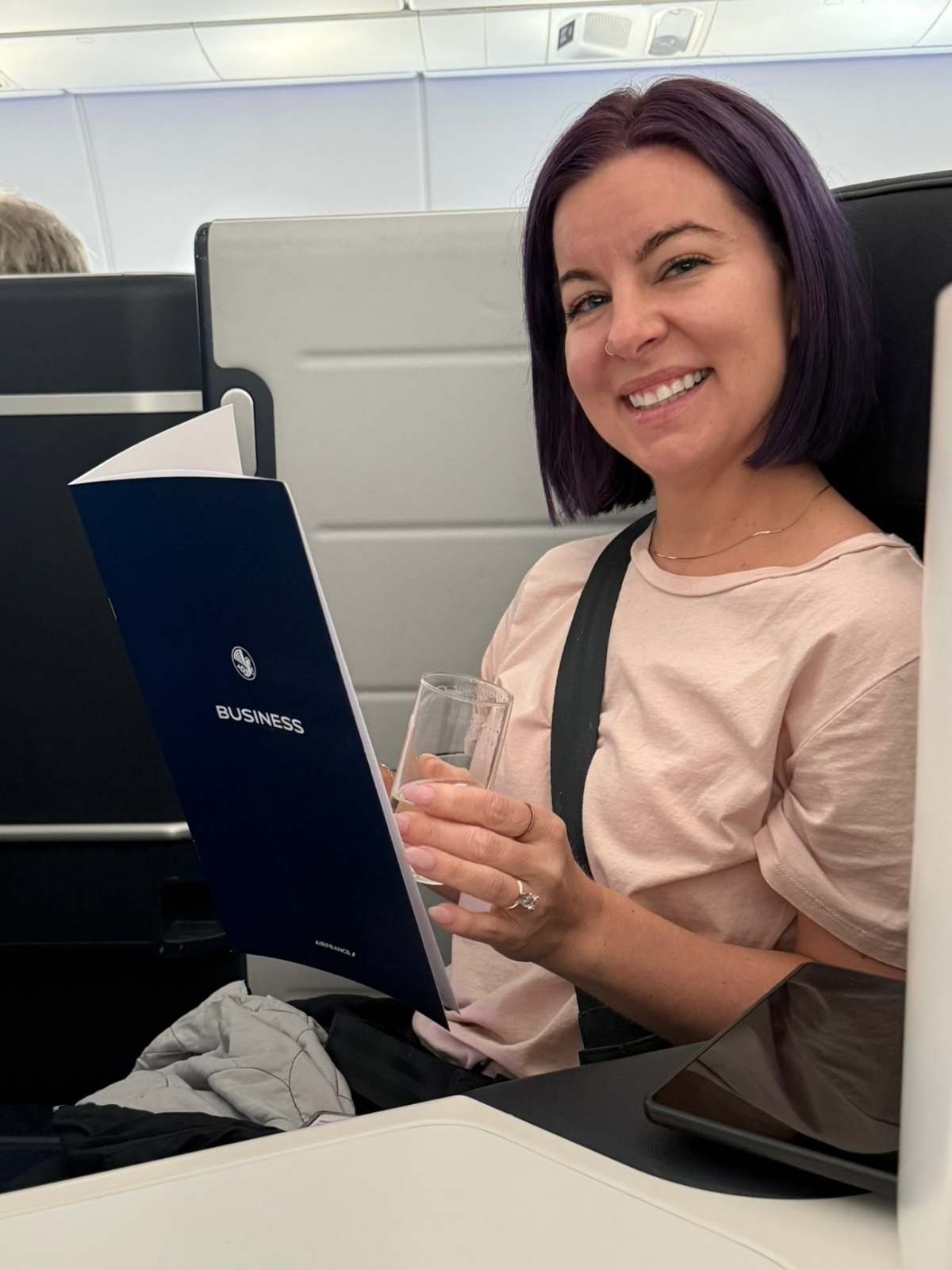 A woman with purple hair smiling on an airplane, holding a glass and a business pamphlet.