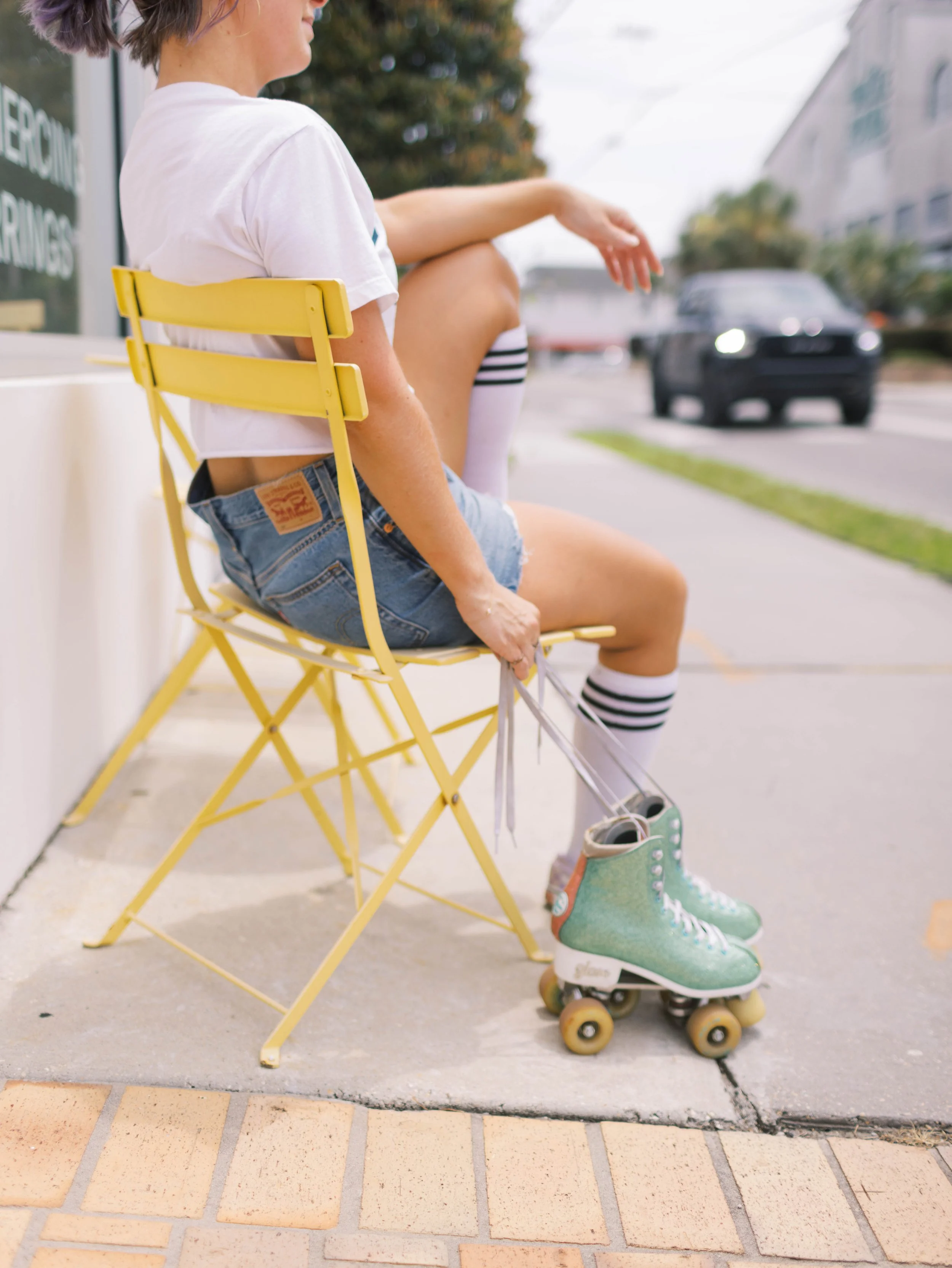 A woman sitting on a yellow foldable chair on a sidewalk, wearing roller skates, striped knee-high socks, denim shorts, a white t-shirt, with a person’s leg stretched out next to her. The background contains buildings, trees, and a parked car, with a partly cloudy sky.
