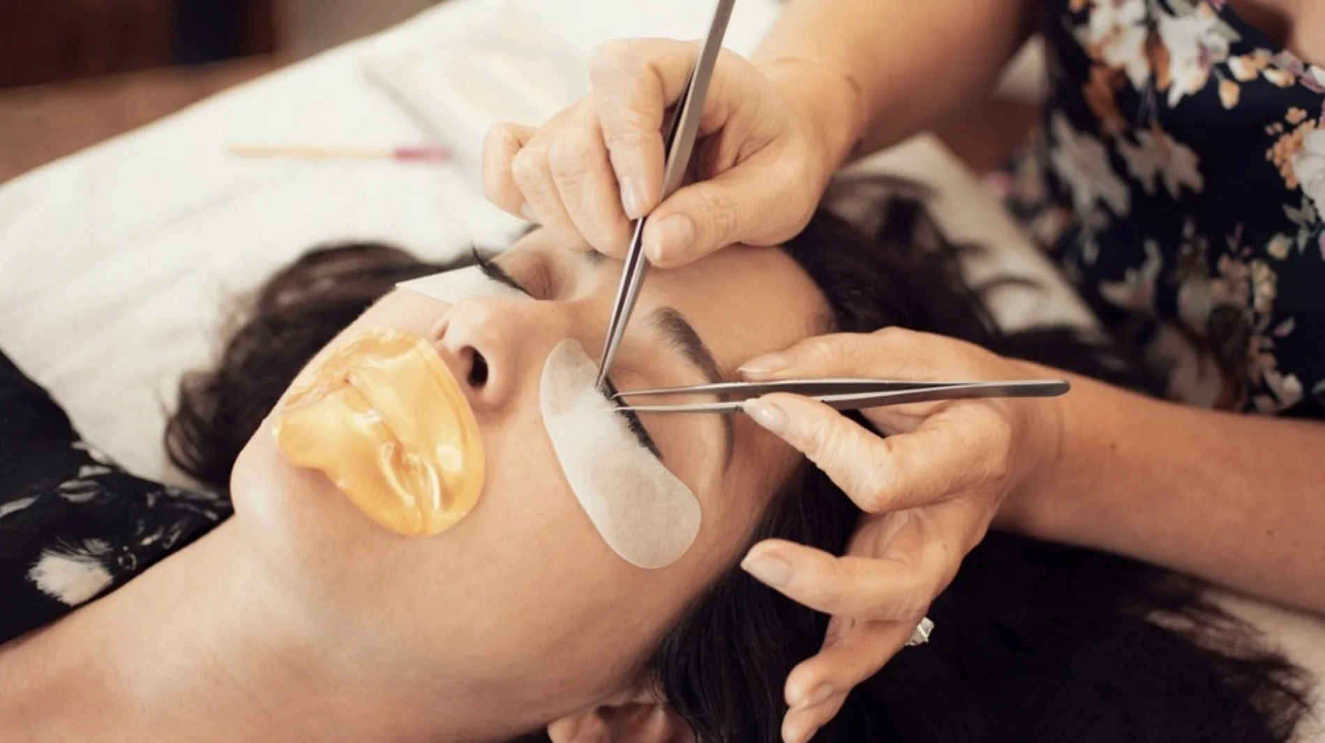 A woman receiving a facial treatment with a gold mask, eye patches, and a patch on her nose while esthetician uses tools near her eyes.