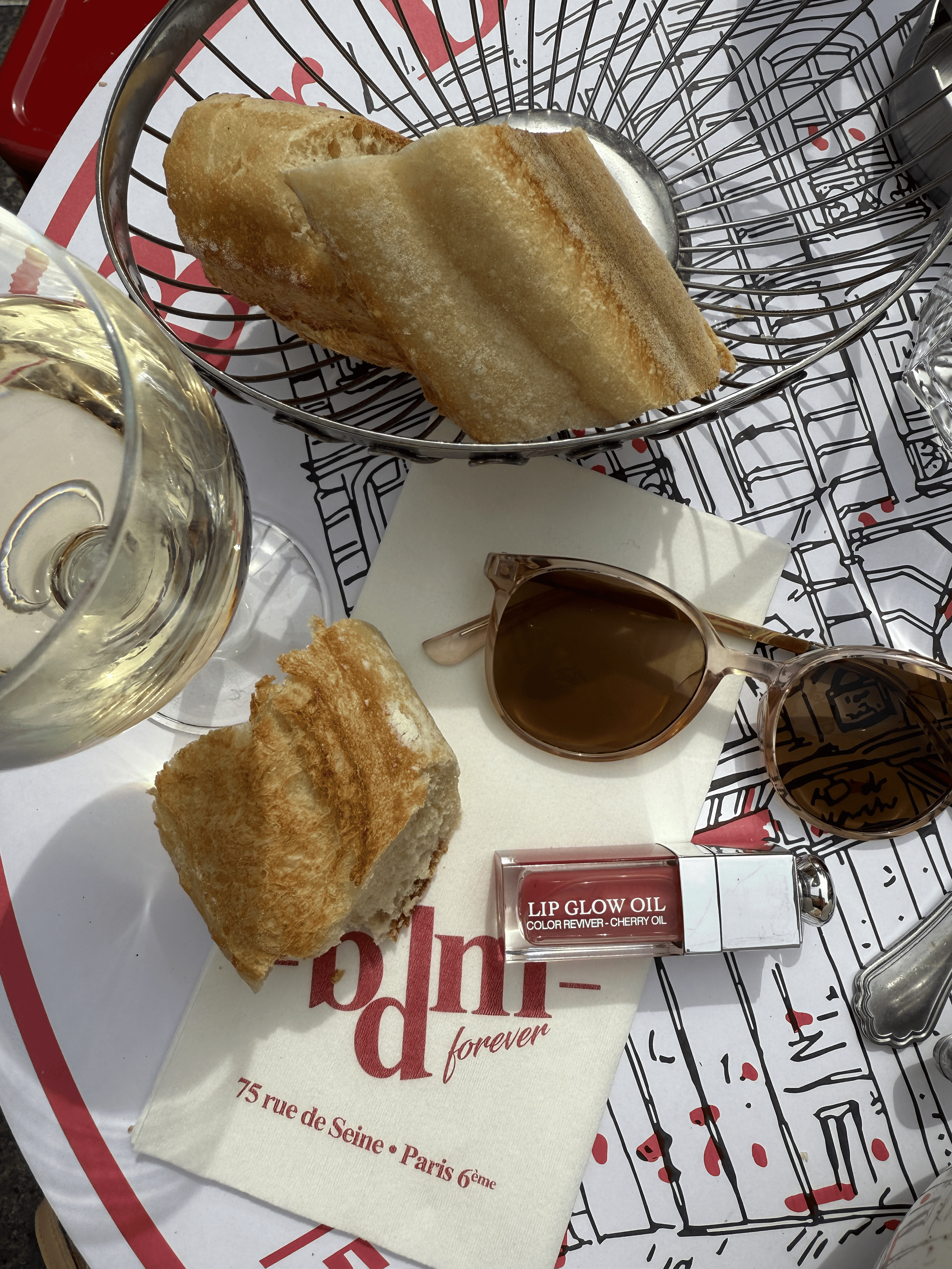 Bread and a glass of white wine on a table with Paris-themed paper and a lip glow oil on a dish.