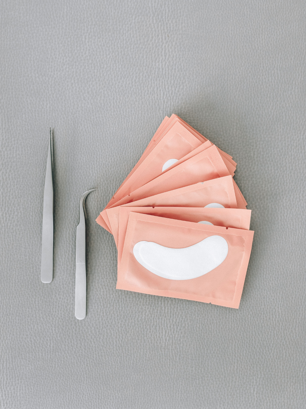 Multiple pink face mask packets with white oval designs, two metal tools with flat and curved ends on a gray textured surface.