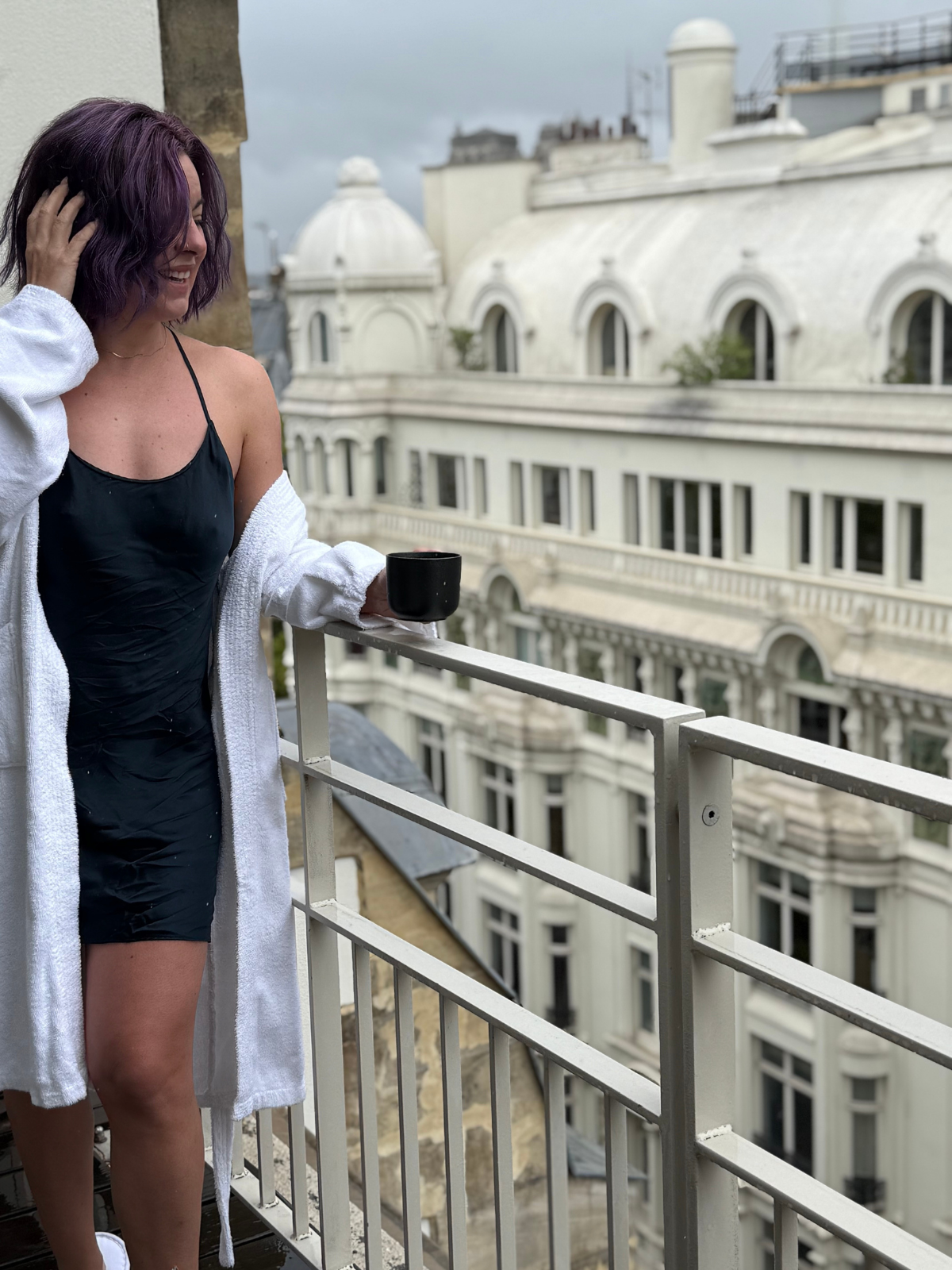 A woman with purple hair, wearing a black slip dress and white bathrobe, standing on a balcony holding a black mug and smiling, with historic buildings in the background under cloudy skies.