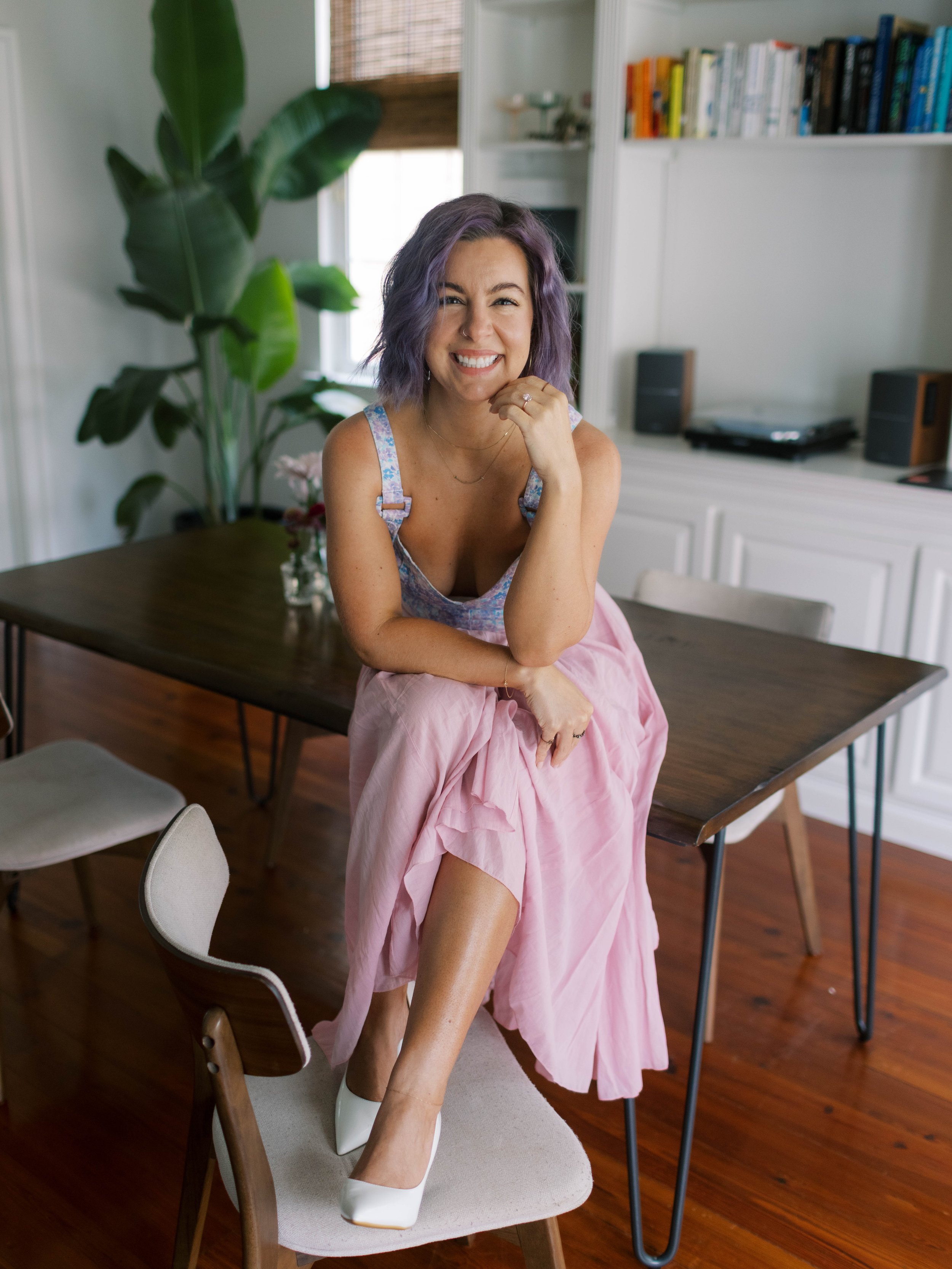 A woman with purple hair smiling and sitting on a table, wearing a floral top and pink skirt, with white shoes, in a room with bookshelves and a large plant in the background.