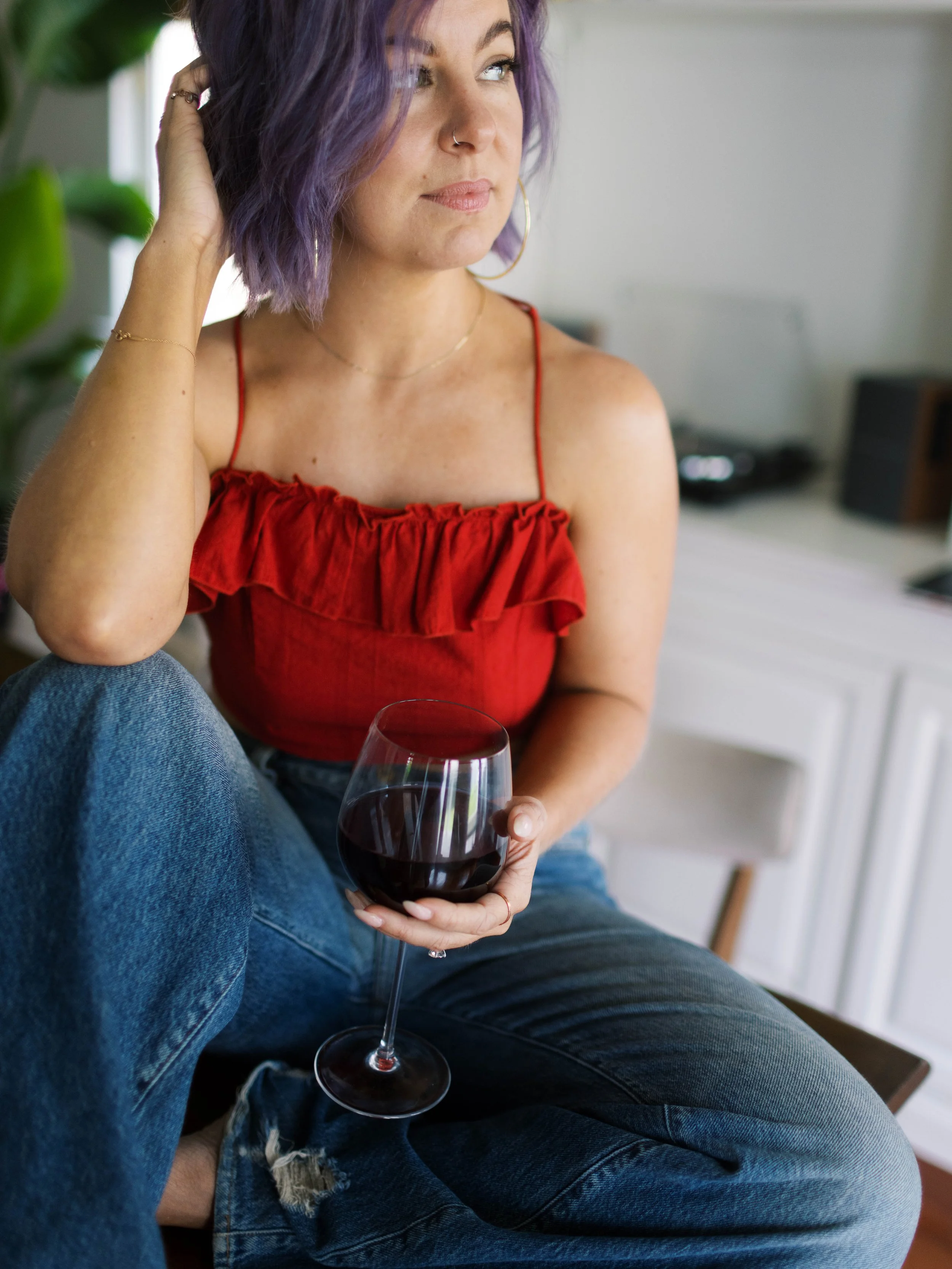 A woman with purple hair, wearing a red top and blue jeans, sitting and holding a glass of red wine.