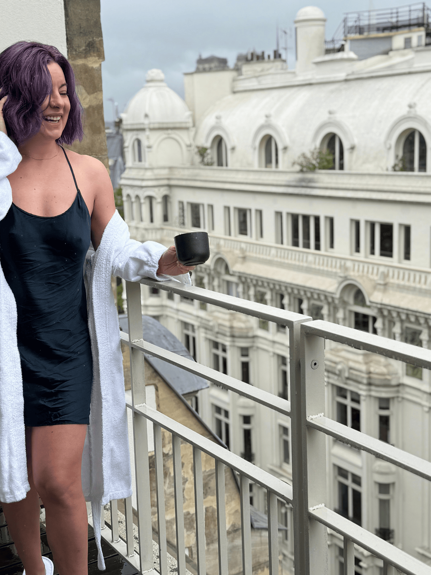 Woman with purple hair wearing a black satin dress and white towel on a balcony in a city with historic architecture.