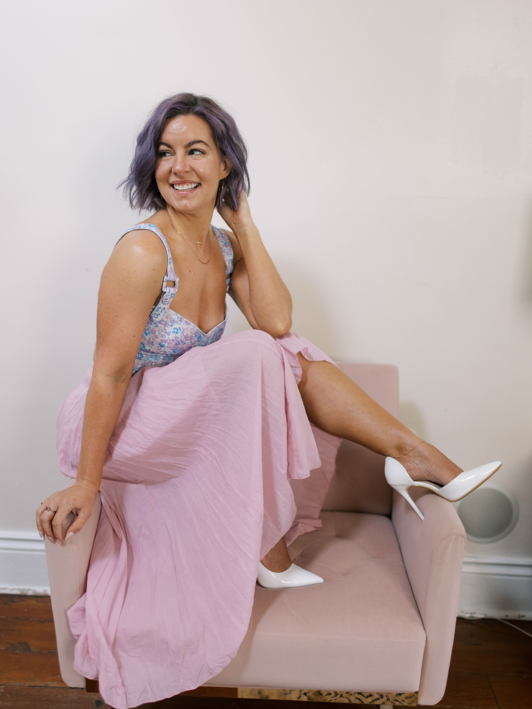 A woman with purple hair sitting on a pink couch, adjusting her loose pink skirt, wearing a floral sleeveless top and white high heels, smiling and looking to the side, against a plain white wall.