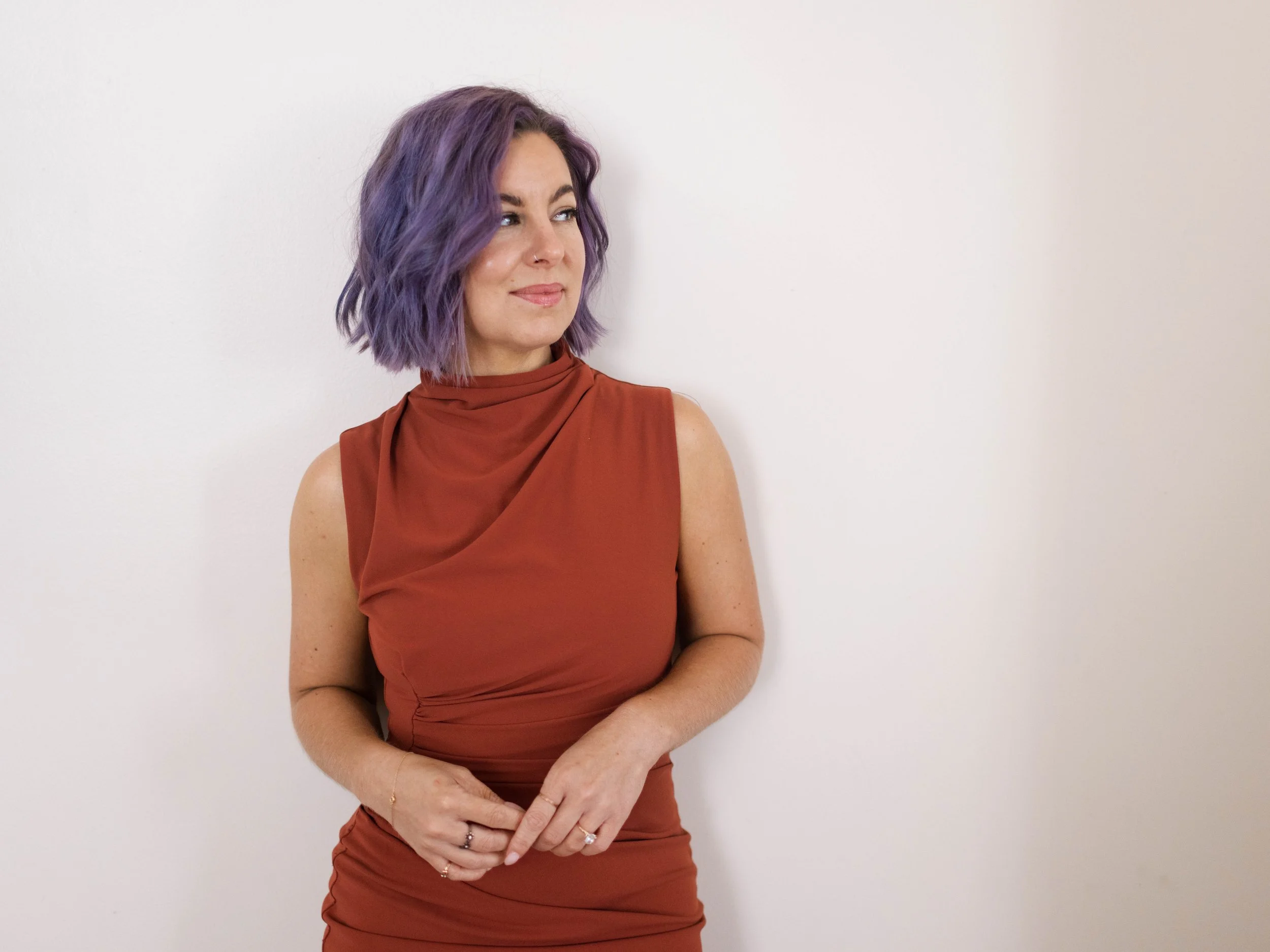 A woman with shoulder-length purple hair, wearing a sleeveless rust-colored dress, is standing against a plain white wall, looking to her right with a slight smile.