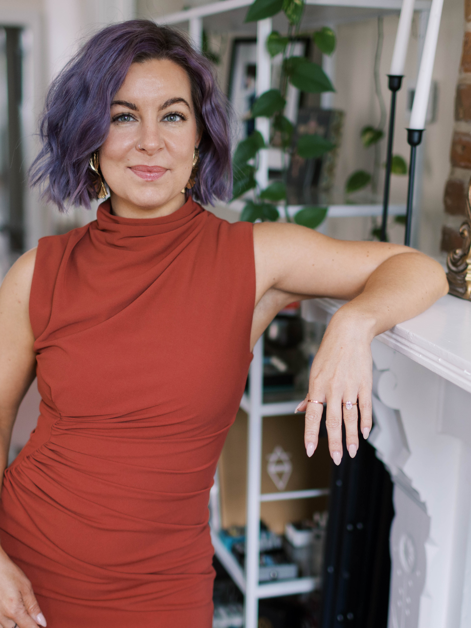 Close-up of a woman with purple hair, wearing a rust-colored sleeveless top, smiling slightly, in a home interior with green plants and decorative items in the background.