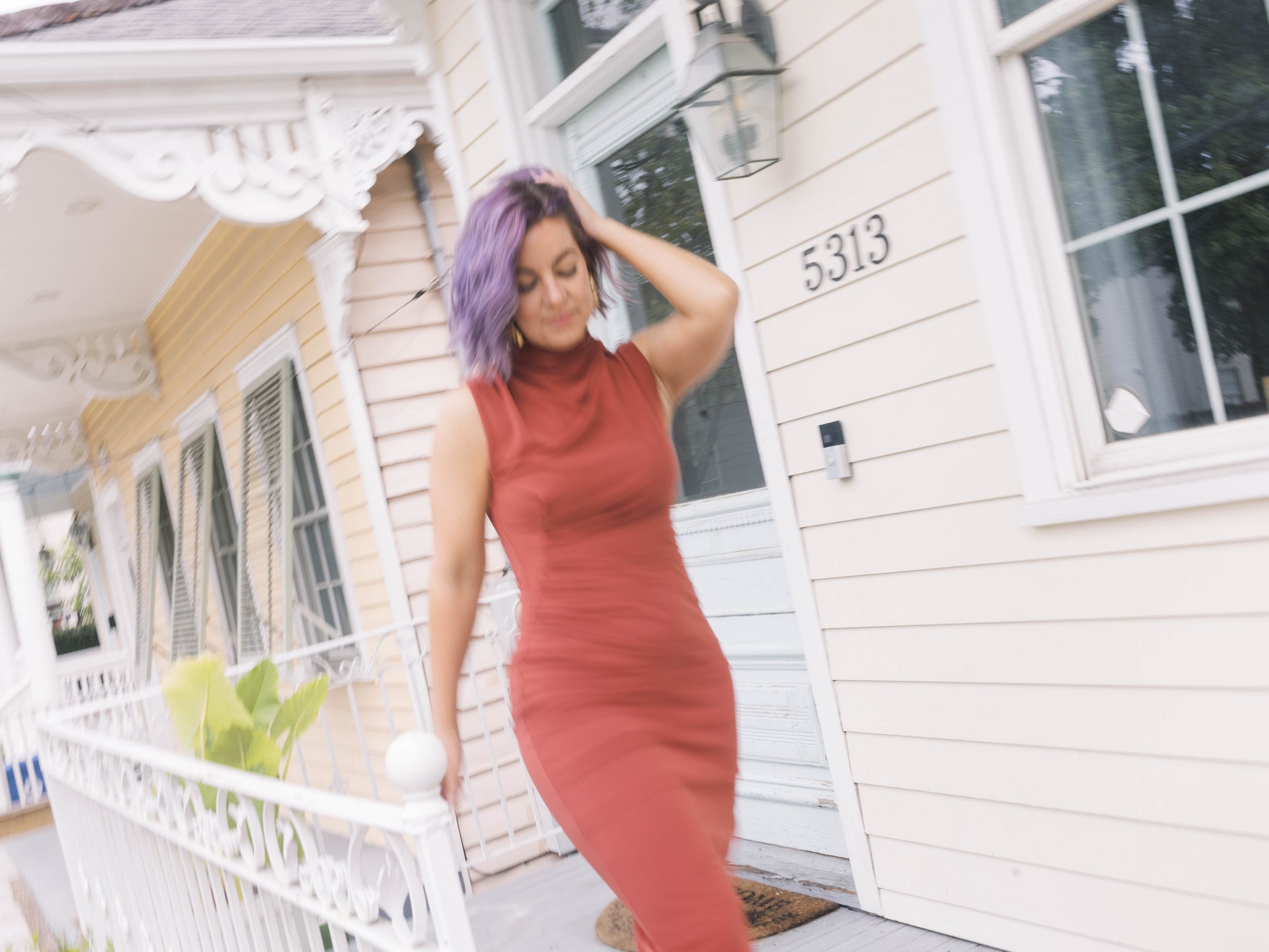 Woman with purple hair in a rust-colored dress resting her hand on her head outside a house with beige siding, white trim, and black house numbers 5313.