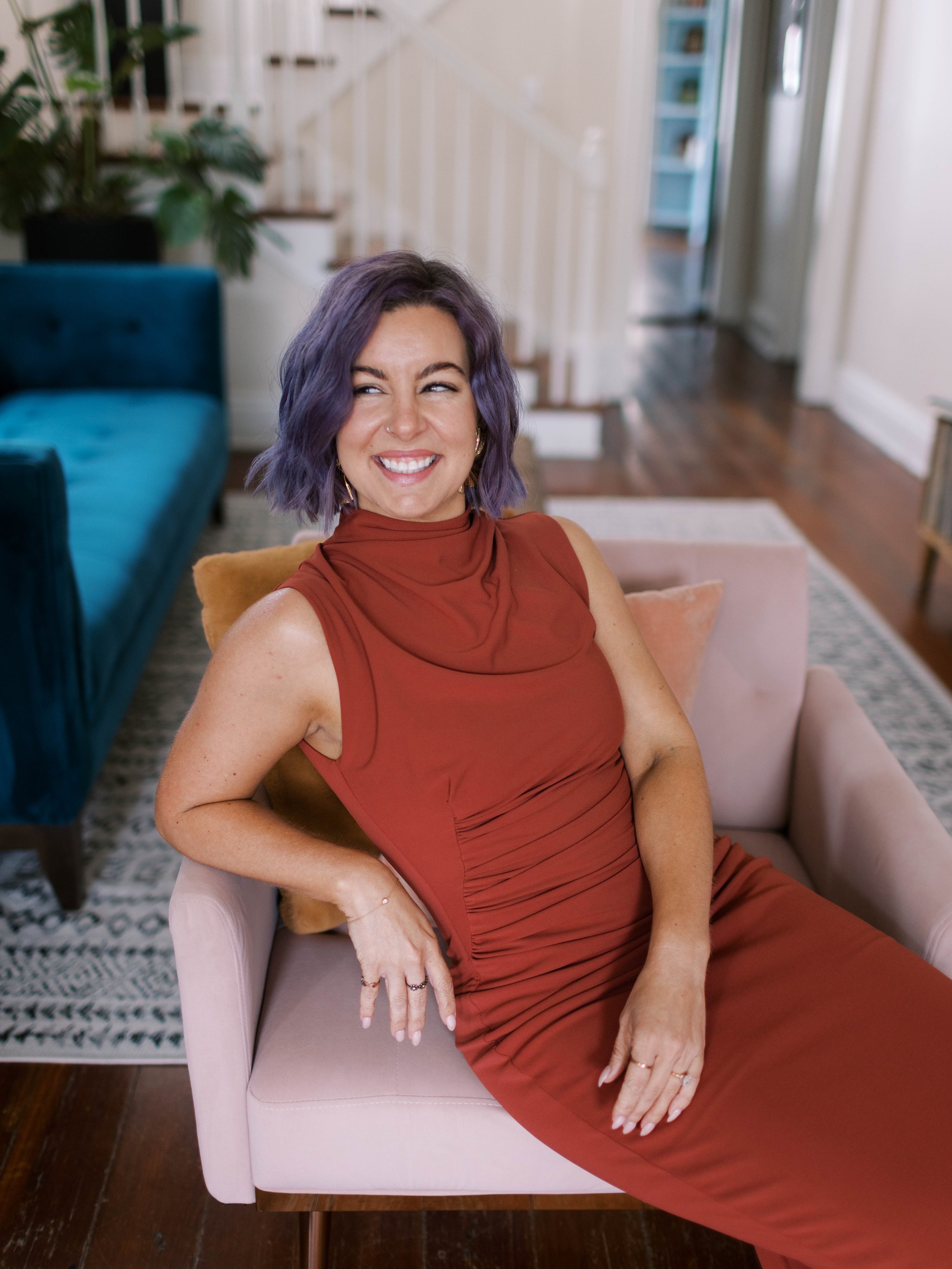 A woman with purple hair and a nose piercing sitting on a pink chair in a cozy living room, smiling and looking to her right.