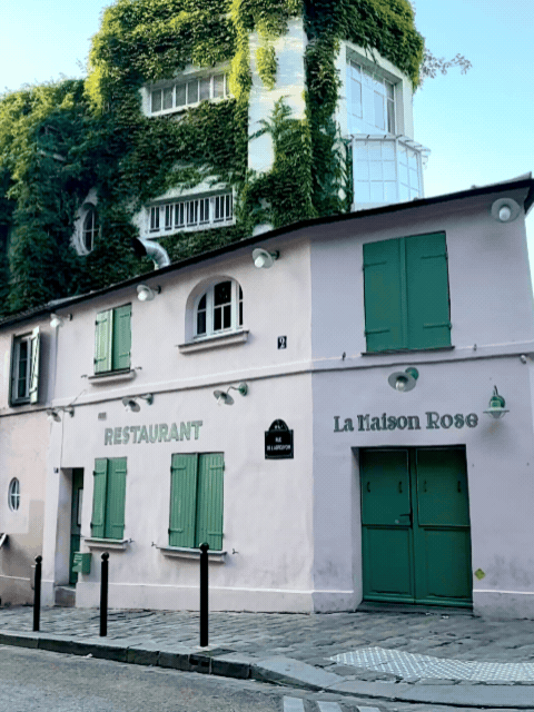 A building with green shutters and a sign that says La Maison Rose, with a large ivy-covered structure behind it.