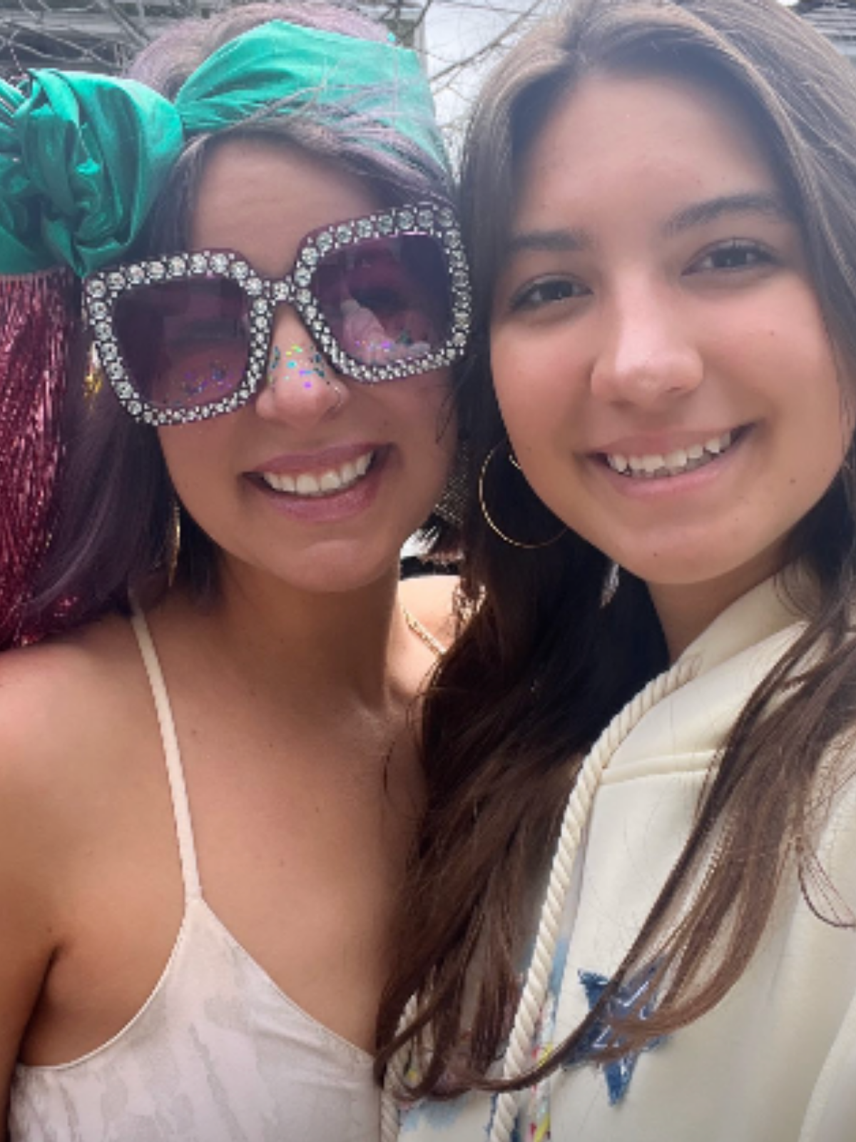 Two women smiling and taking a close-up selfie outdoors, one wearing large heart-shaped sunglasses decorated with rhinestones, a green headscarf, and a white top with spaghetti straps, and the other with long brown hair, hoop earrings, and a cream-colored hoodie.
