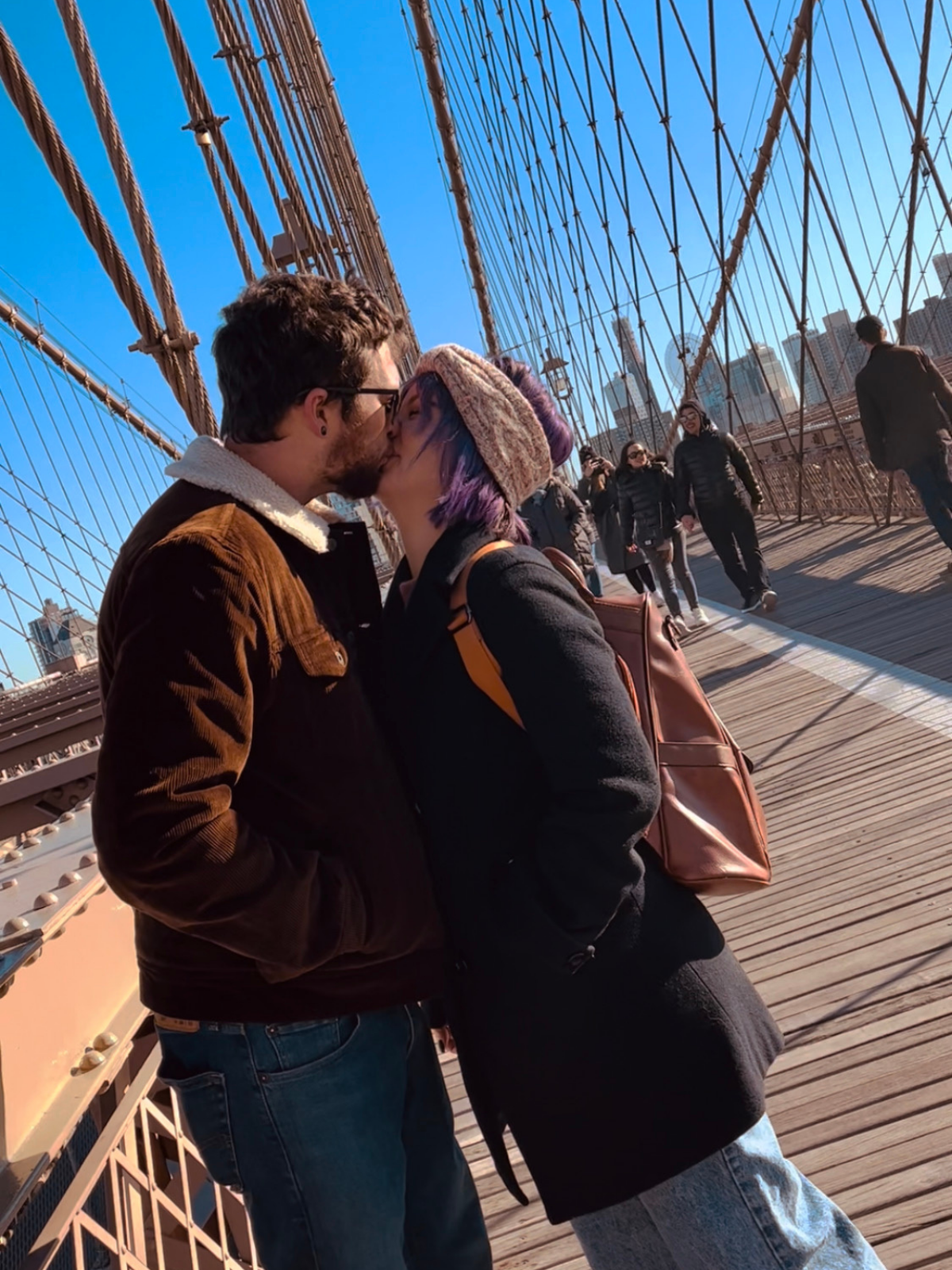 A couple sharing a kiss on the Brooklyn Bridge with pedestrians walking in the background and New York City skyline visible.