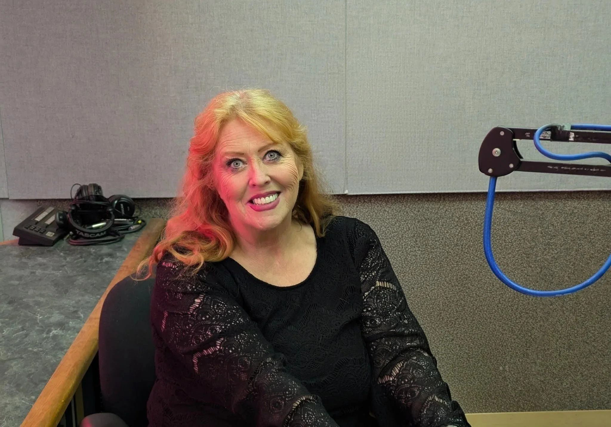 A smiling woman with red hair and blue eyes sitting in a recording studio. There are headphones and audio equipment on the table next to her, and a gray soundproof wall behind her.