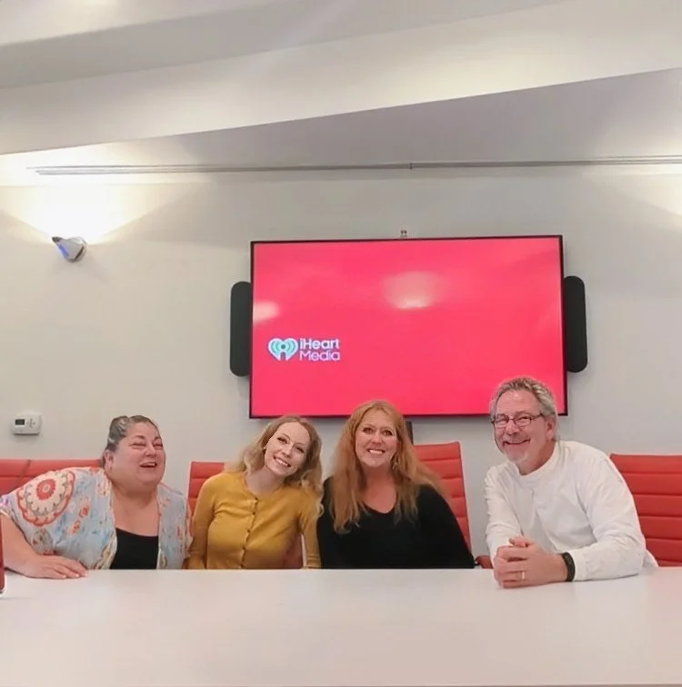 Four people sitting at a conference table, smiling, with a large screen behind them displaying the iHeart Media logo.