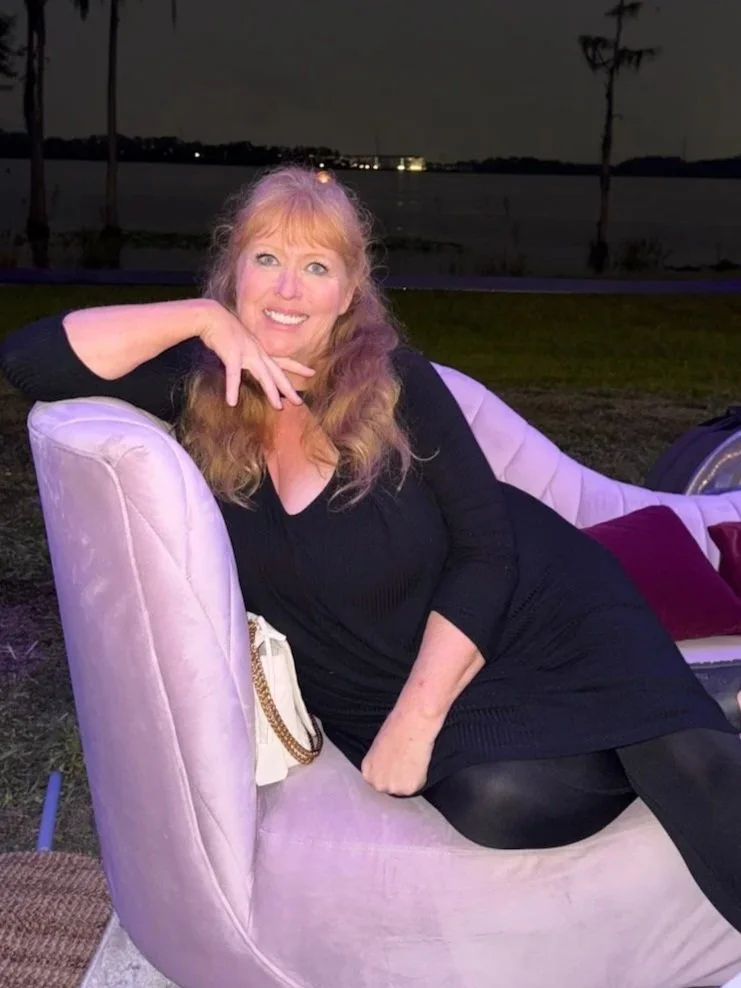 A woman with long, blonde hair is lounging on a pink velvet couch outdoors at night. She is smiling, resting her head on her left hand, and wearing a black dress with leggings. The background shows a dark sky, some trees, and a body of water with city lights reflecting on it.