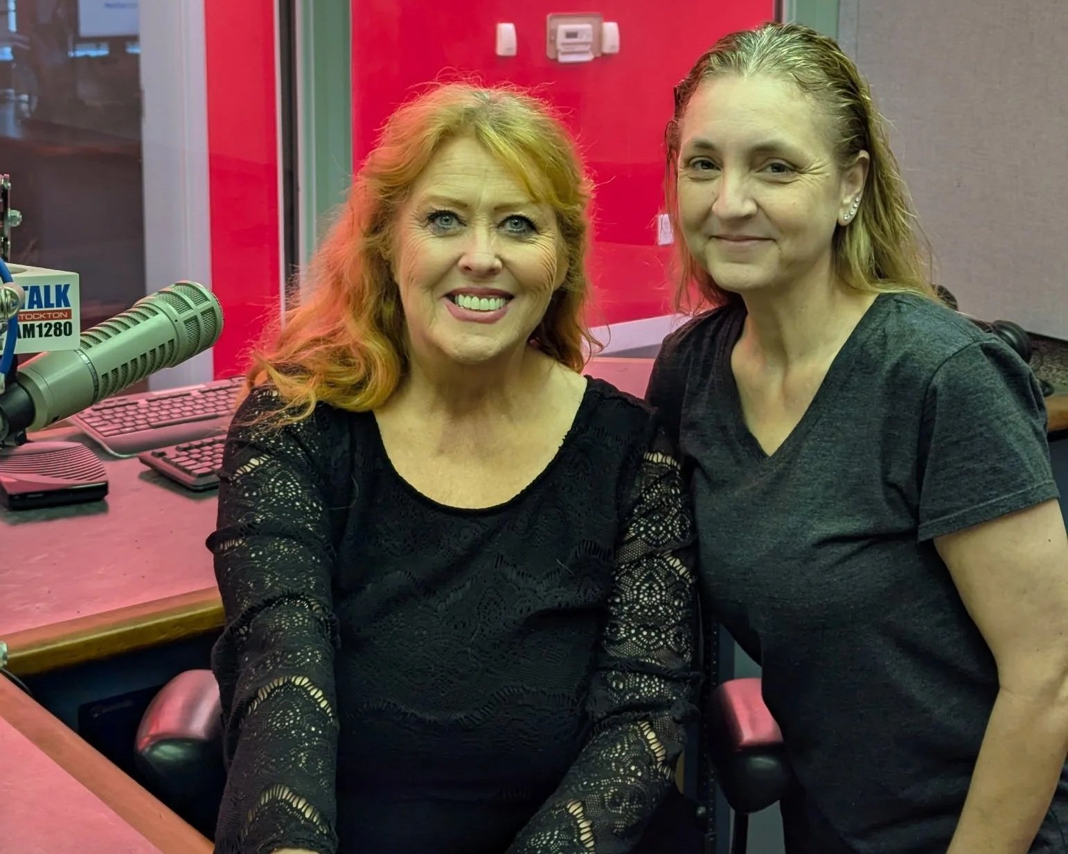 Two women sitting in a radio broadcasting studio. One woman has red hair and is smiling, the other has blonde hair and is smiling softly. There is radio equipment in front of them, including a microphone and a computer keyboard.