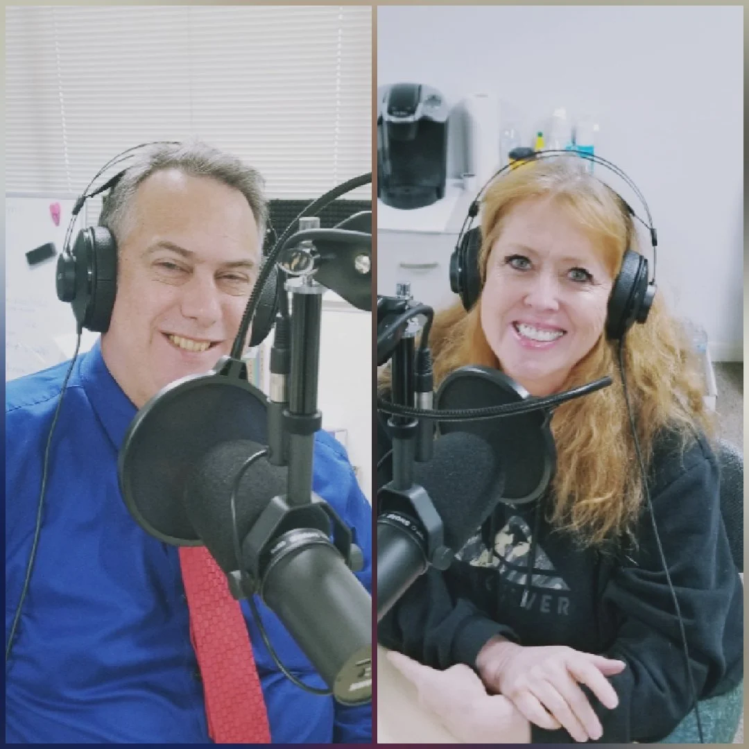 Two people in a recording studio, wearing headphones and speaking into microphones. The man on the left has gray hair and is smiling, wearing a blue shirt. The woman on the right has red hair and is smiling, wearing a black sweatshirt.