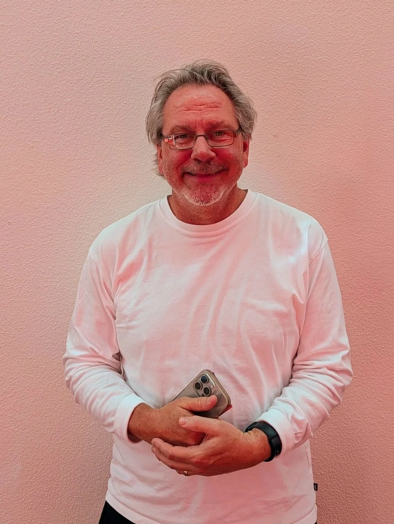 A smiling middle-aged man with glasses and gray hair, wearing a white long-sleeve shirt, holding a smartphone, standing against a light pink textured wall.