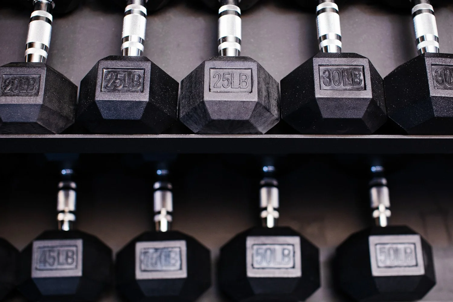 Two rows of dumbbells with silver handles. The top row has three 25-pound dumbbells and two 30-pound dumbbells, while the bottom row has three 45-pound, 75-pound, and 50-pound dumbbells. Atlanta Indoor Golf simulator and strength training facility.