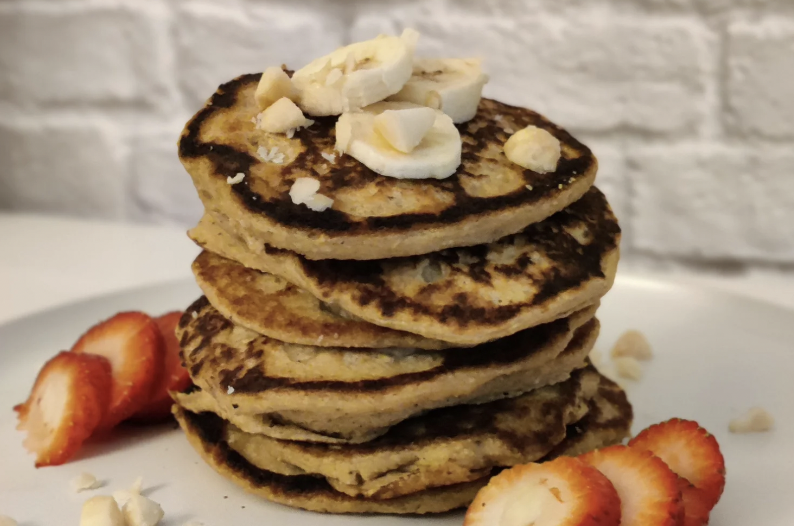 A stack of plantain-oat pancakes on a white plate surrounded by cut strawberries with a few slices of plantain on top