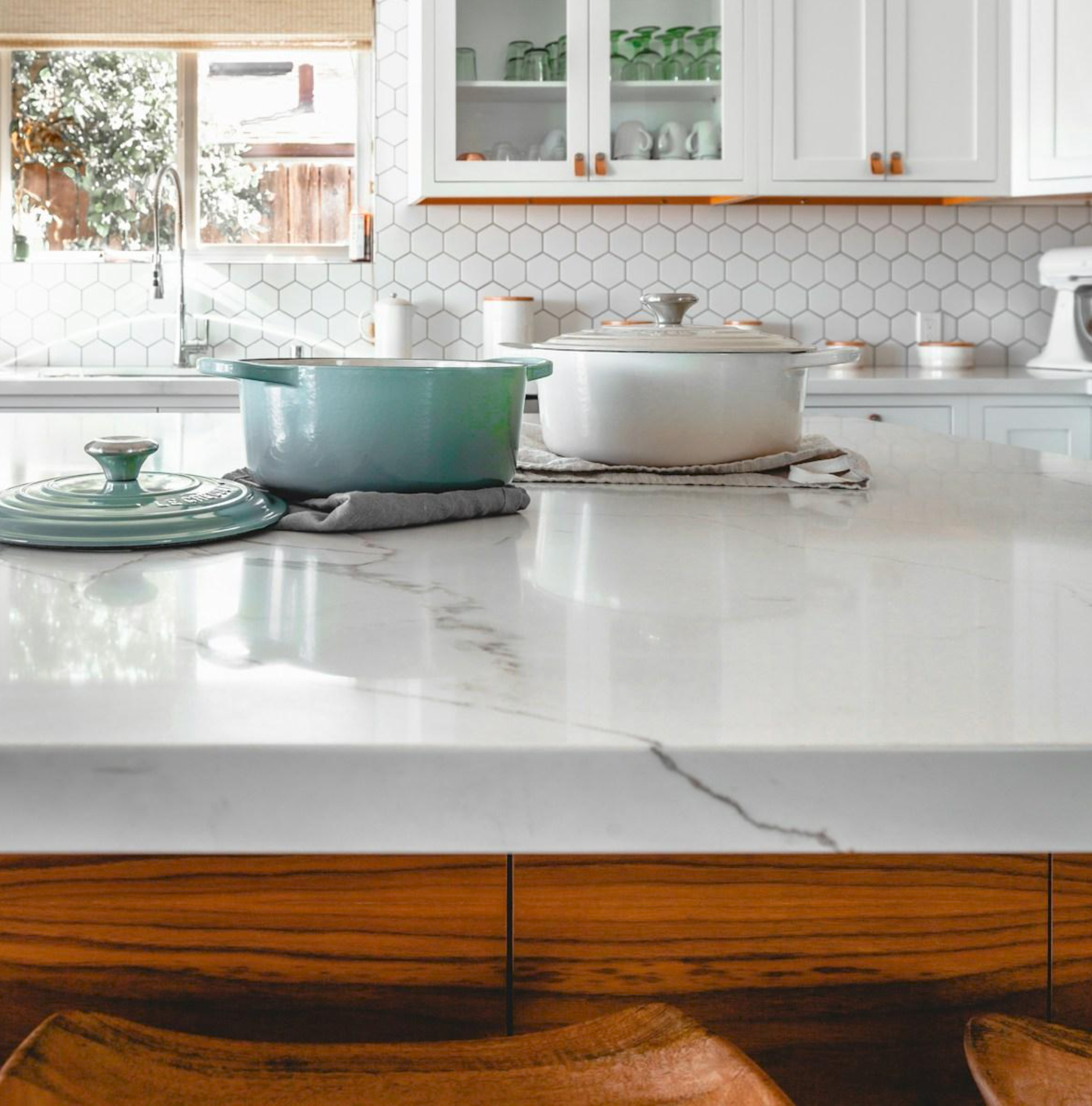 Two dutch ovens, one lidless sea foam green and one white with a lid, on a white marble kitchen counter with white tiles and cabinets in the background