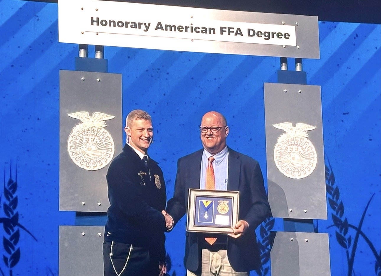 Mike Armbruster shaking hands at an award ceremony, one in a police uniform and the other in a suit holding a framed display, with a blue backdrop and FFA emblems, under a sign that reads "Honorary American FFA Degree."