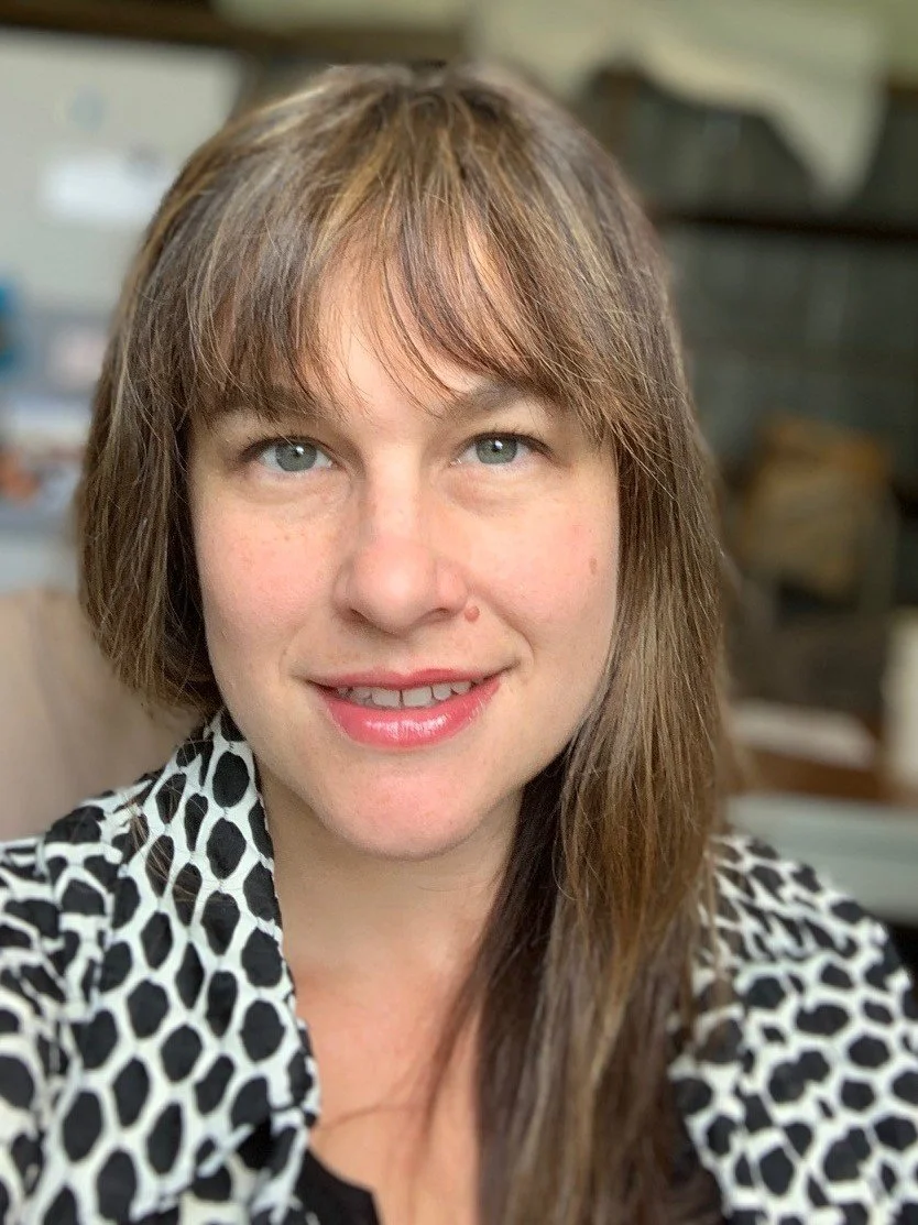 A woman with shoulder-length brown hair and blue eyes, smiling while wearing a black and white polka dot top, sitting indoors.