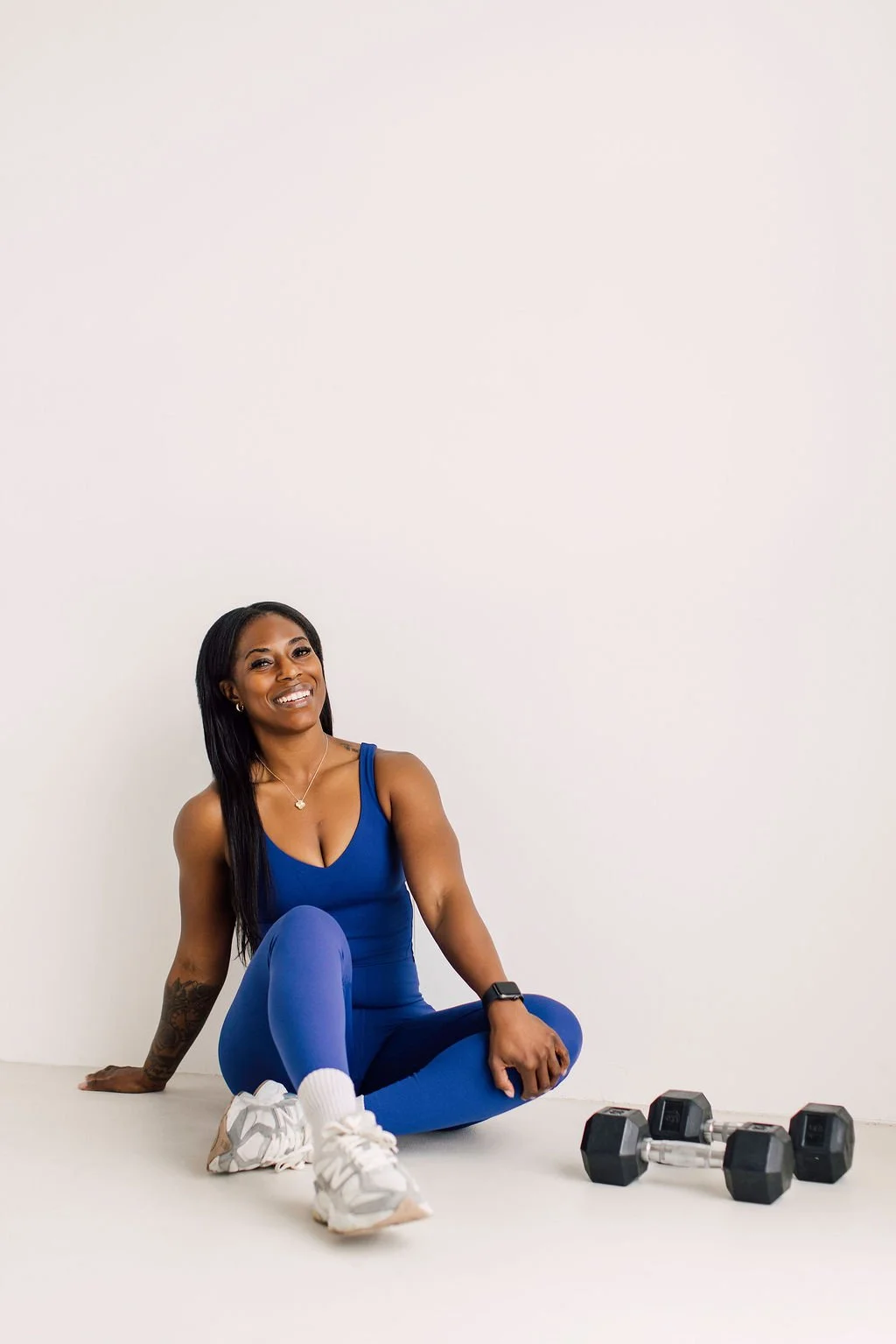 A woman in blue workout clothing sitting on the floor with dumbbells and a fitness tracker, smiling at the camera.