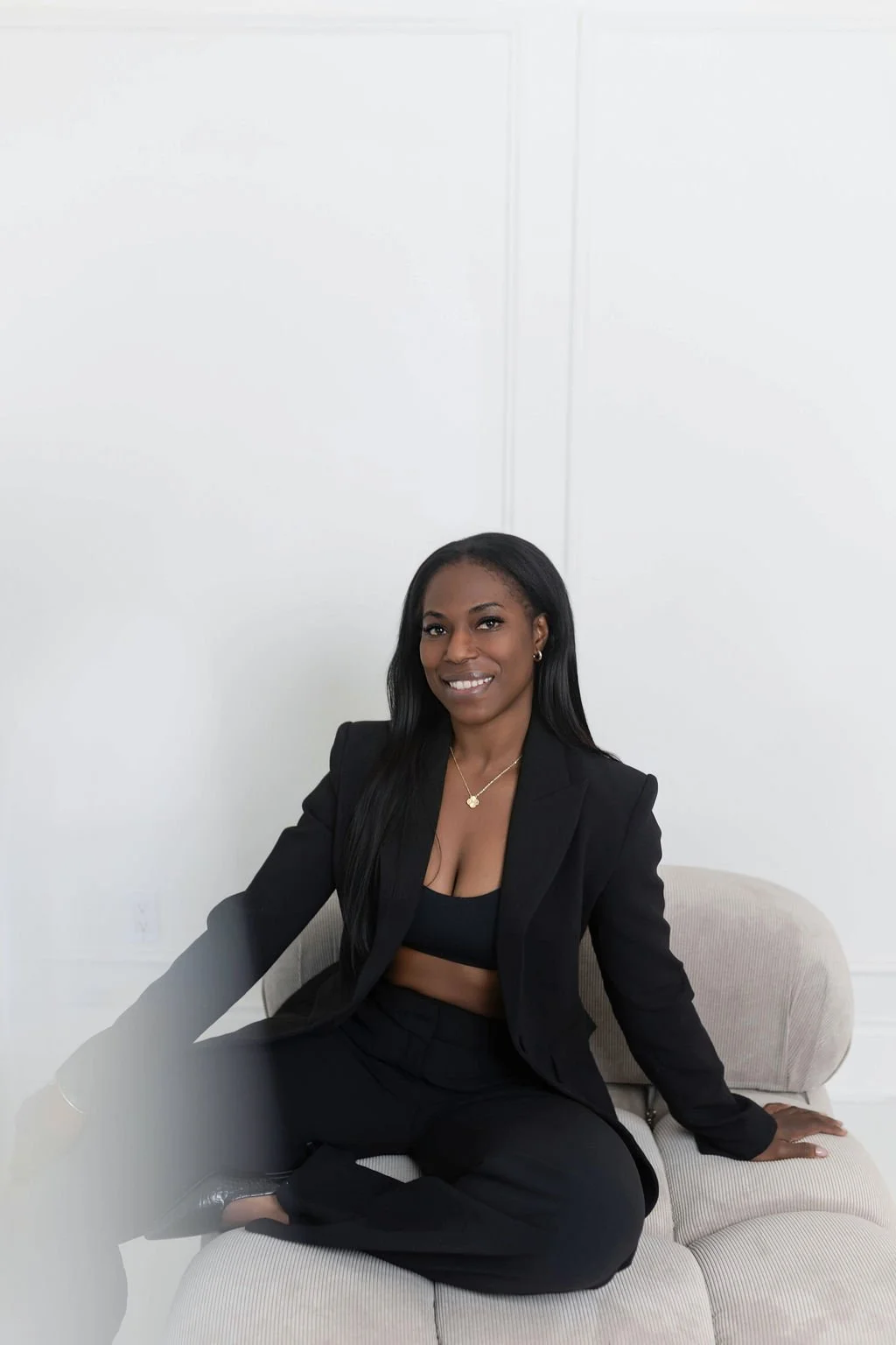 A woman in a black blazer and top, black pants, sitting on a beige couch against a white wall, smiling at the camera.