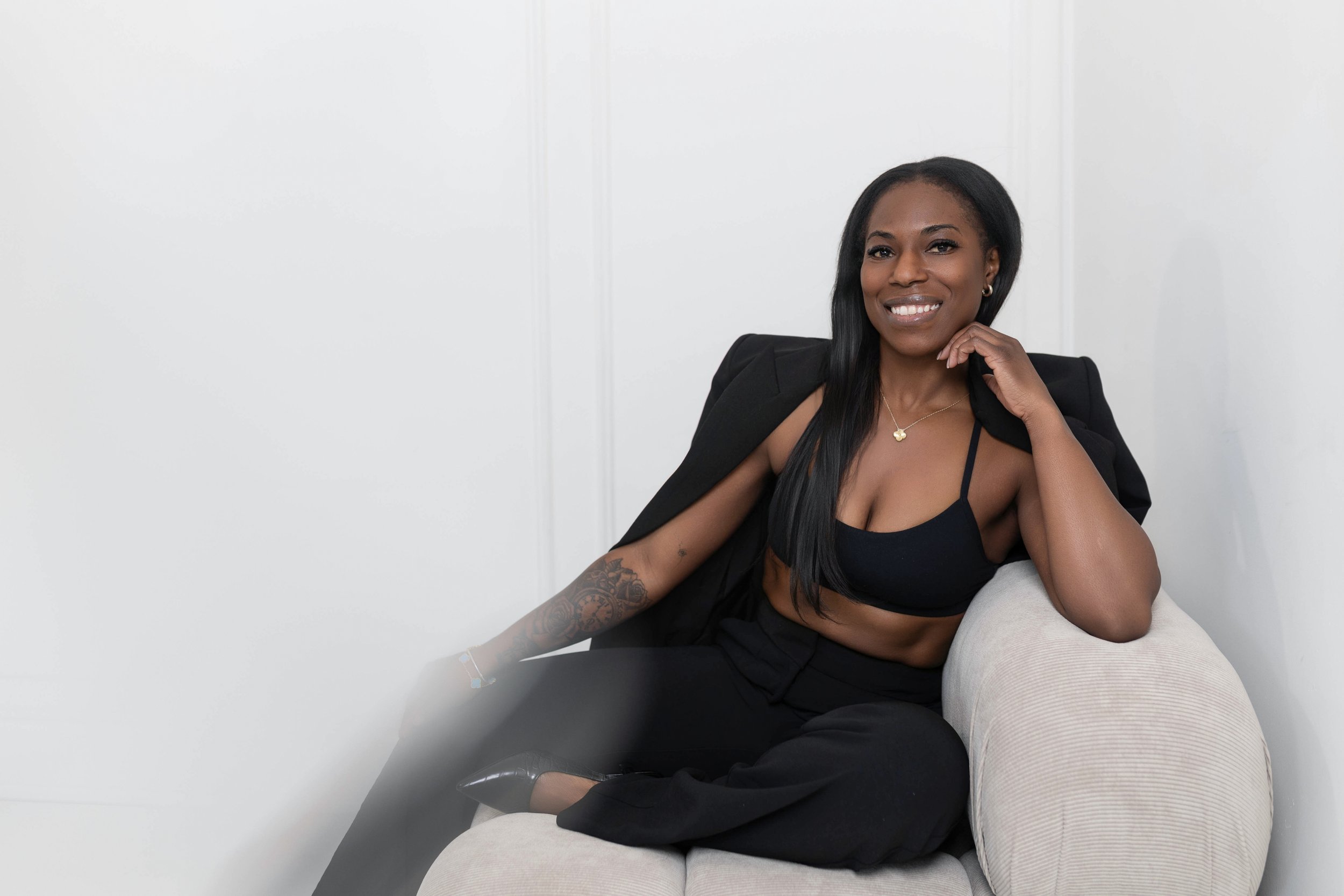 A woman sitting on a beige sofa, smiling, wearing a black, strappy crop top, black pants, and a black blazer draped over her shoulders, in a white room.