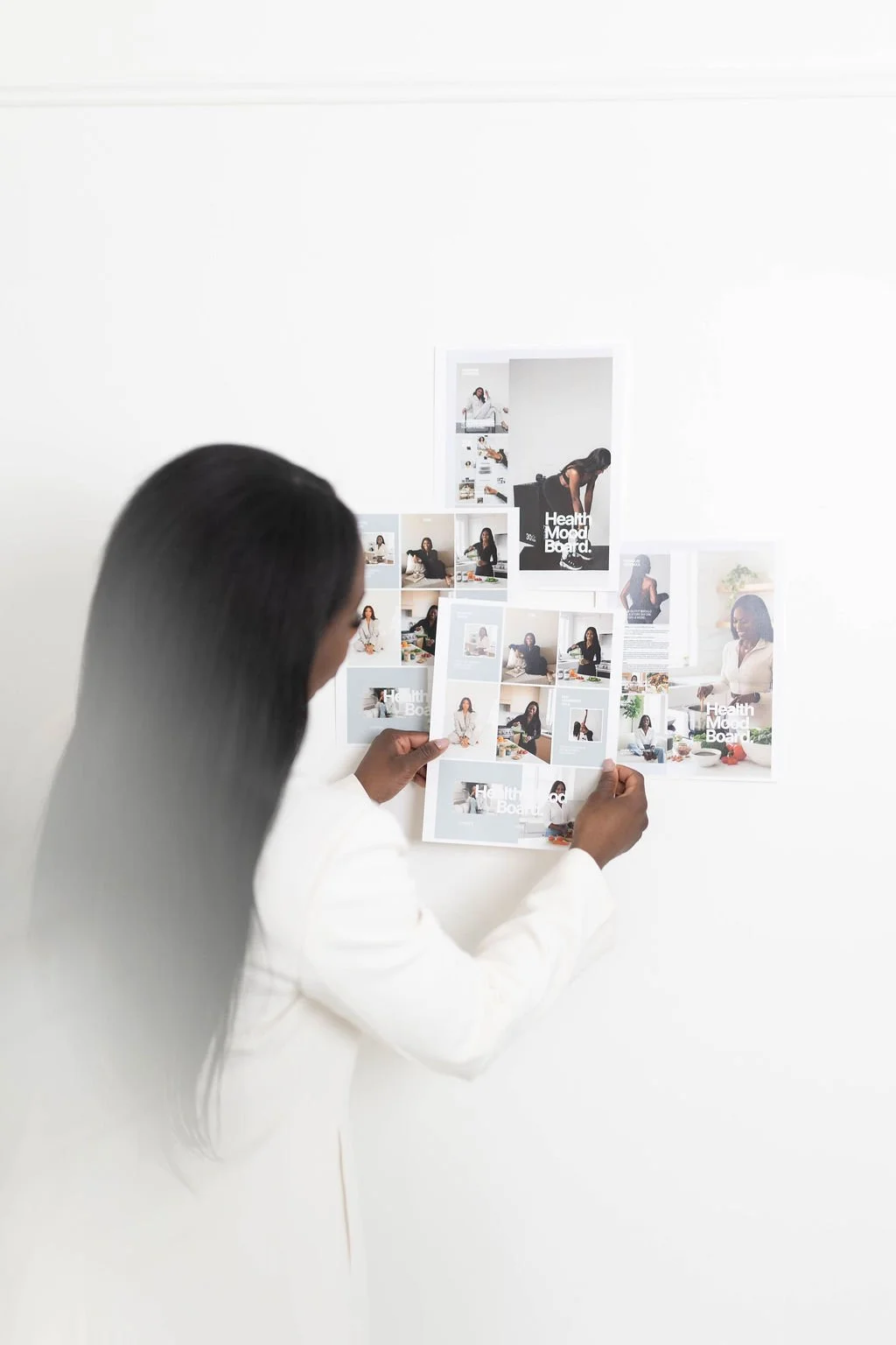A woman in a white blazer with long dark hair looks at a collage of photos and a magazine or flyer on a white wall.