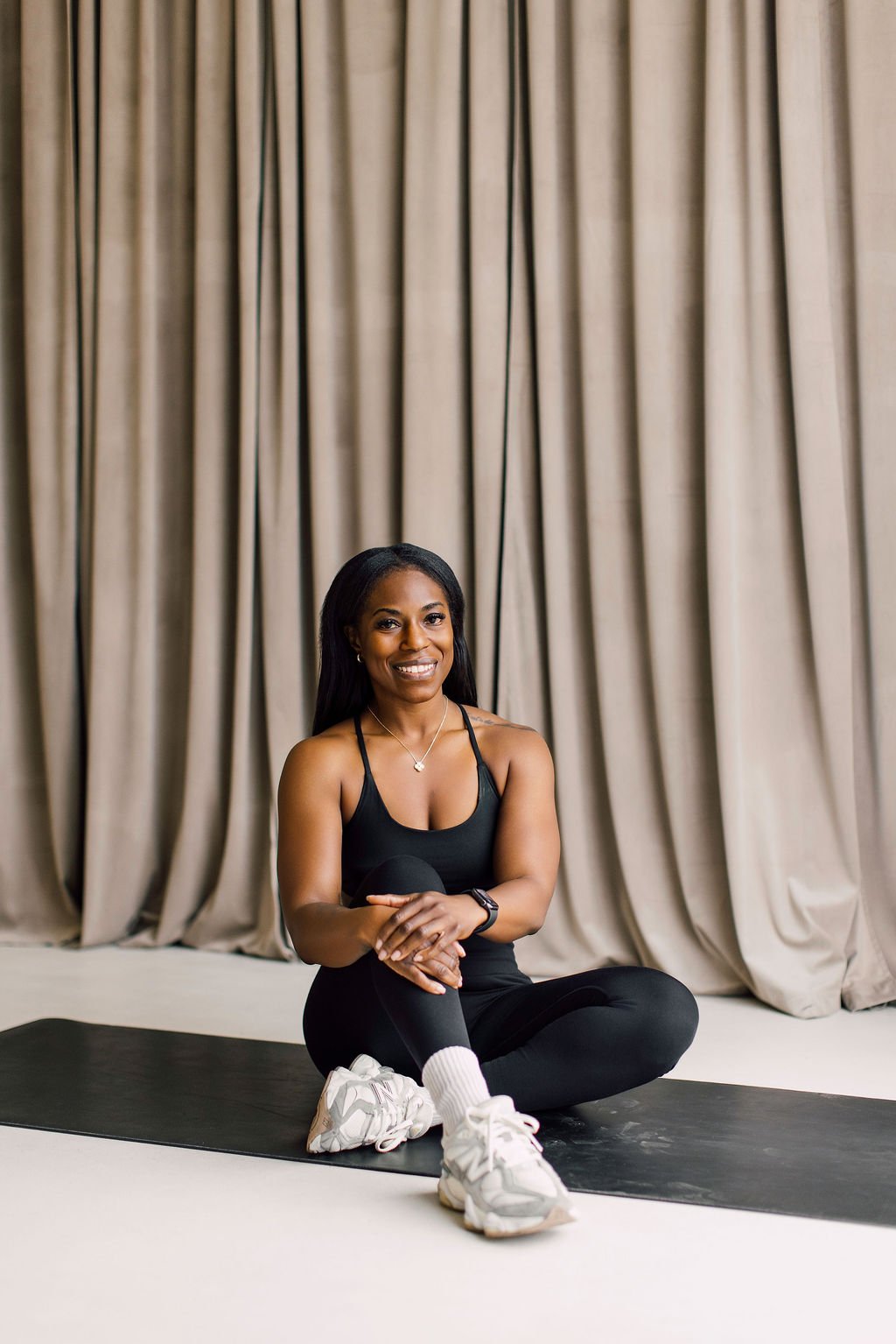 A woman sitting cross-legged on a yoga mat in front of beige curtains, smiling, wearing black workout clothes and white sneakers.