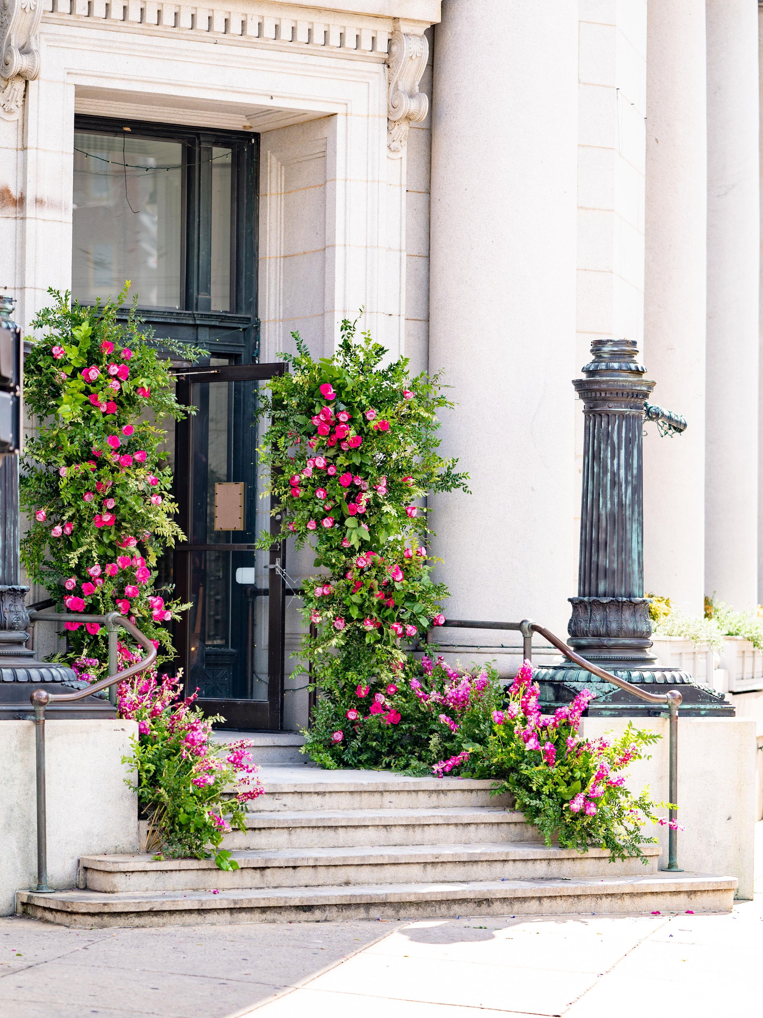 Stone steps leading to an arched doorway with black trim, decorated with pink and purple flowers on either side and around the railing, with classical columns nearby.