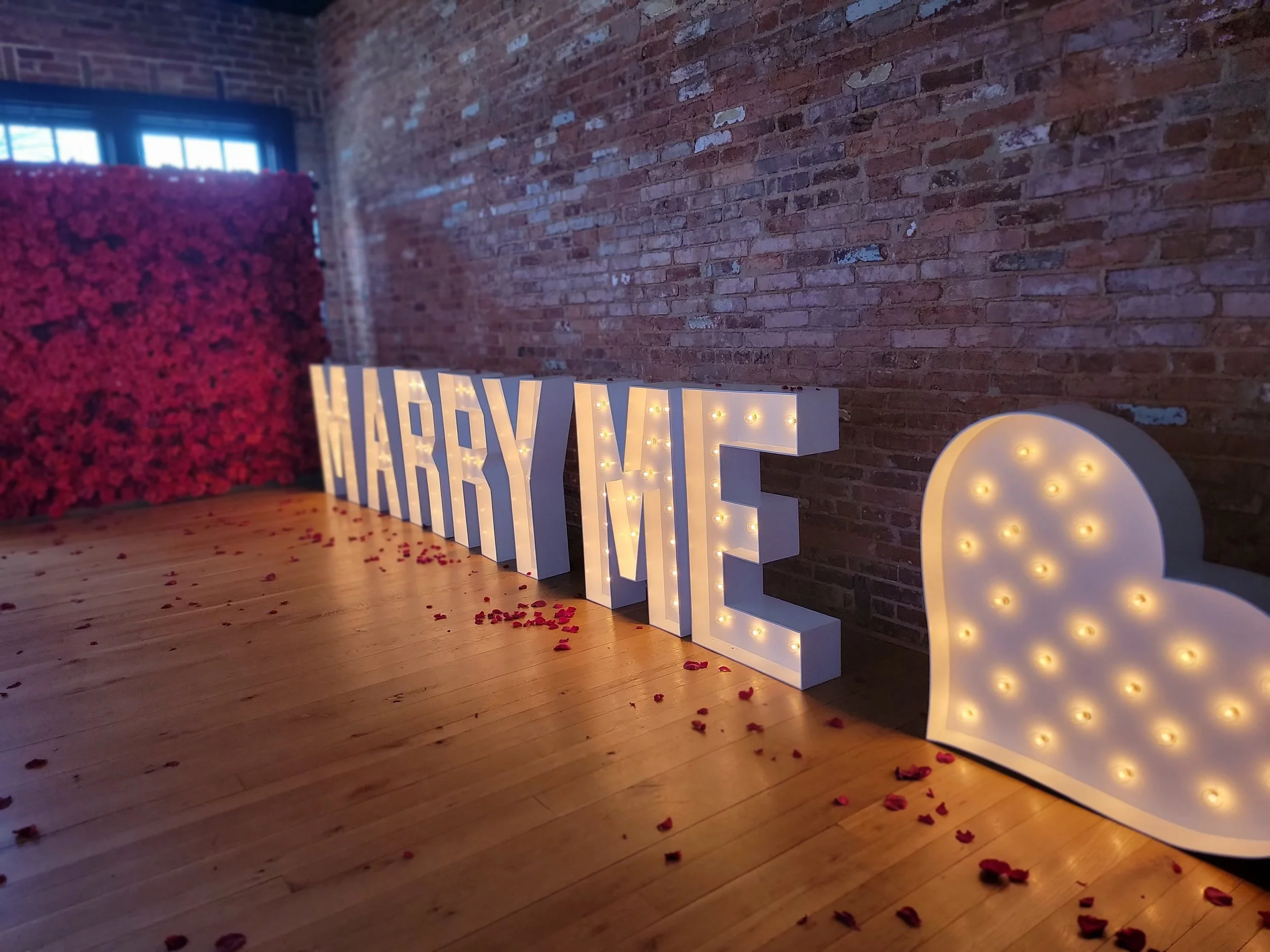 Light-up marquee letters spelling 'HAPPY M' next to a glowing cloud-shaped sign, with rose petals scattered on a wooden floor against a brick wall backdrop.