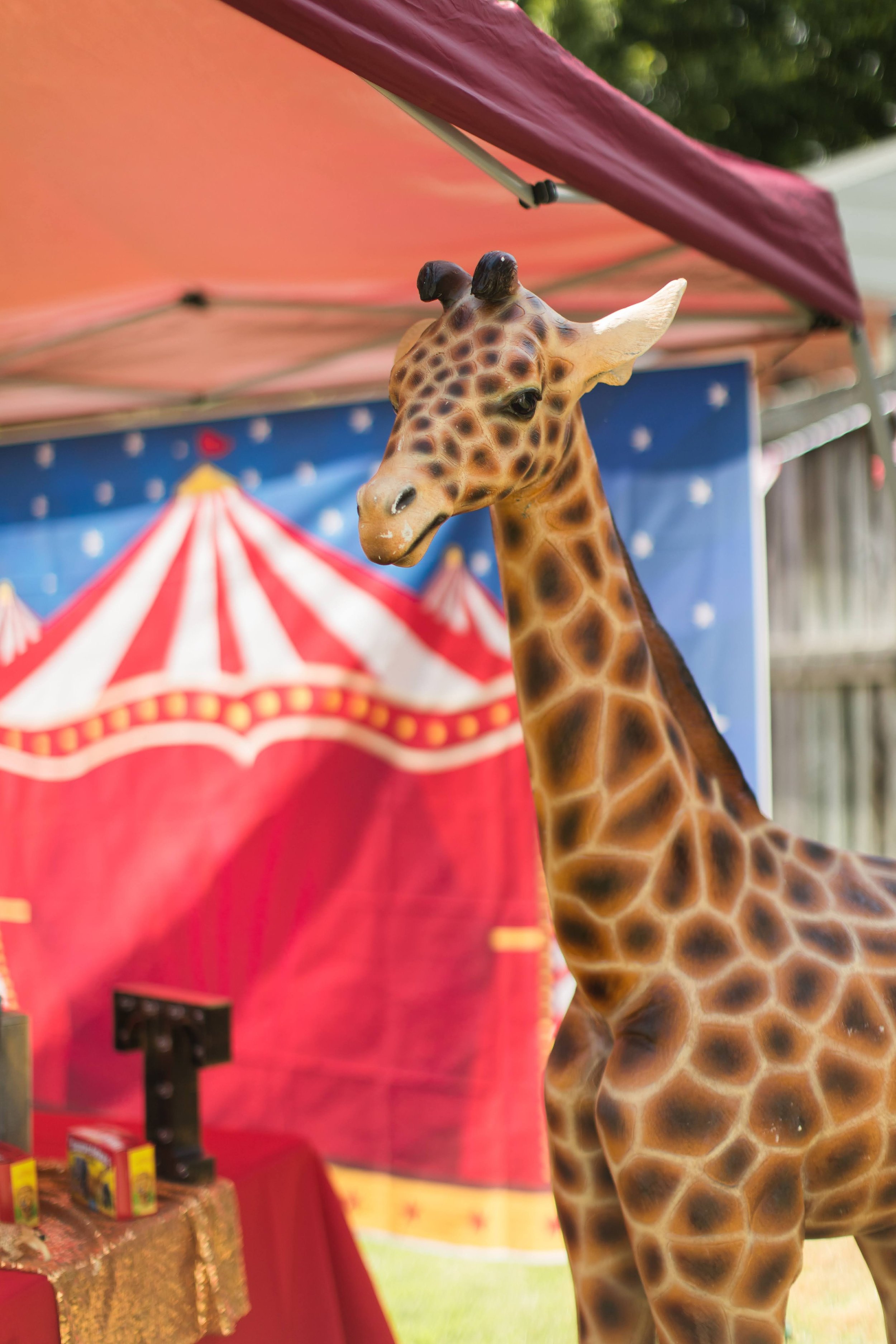 A realistic-looking giraffe statue in front of a circus-themed backdrop and tent. The scene is outdoors with trees and a fence visible in the background.