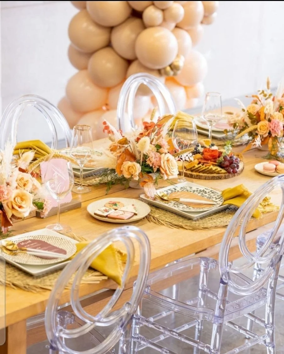 Elegant dining table decorated with floral centerpieces, plates, glasses, and napkins, with a balloon backdrop of beige and peach-colored balloons.