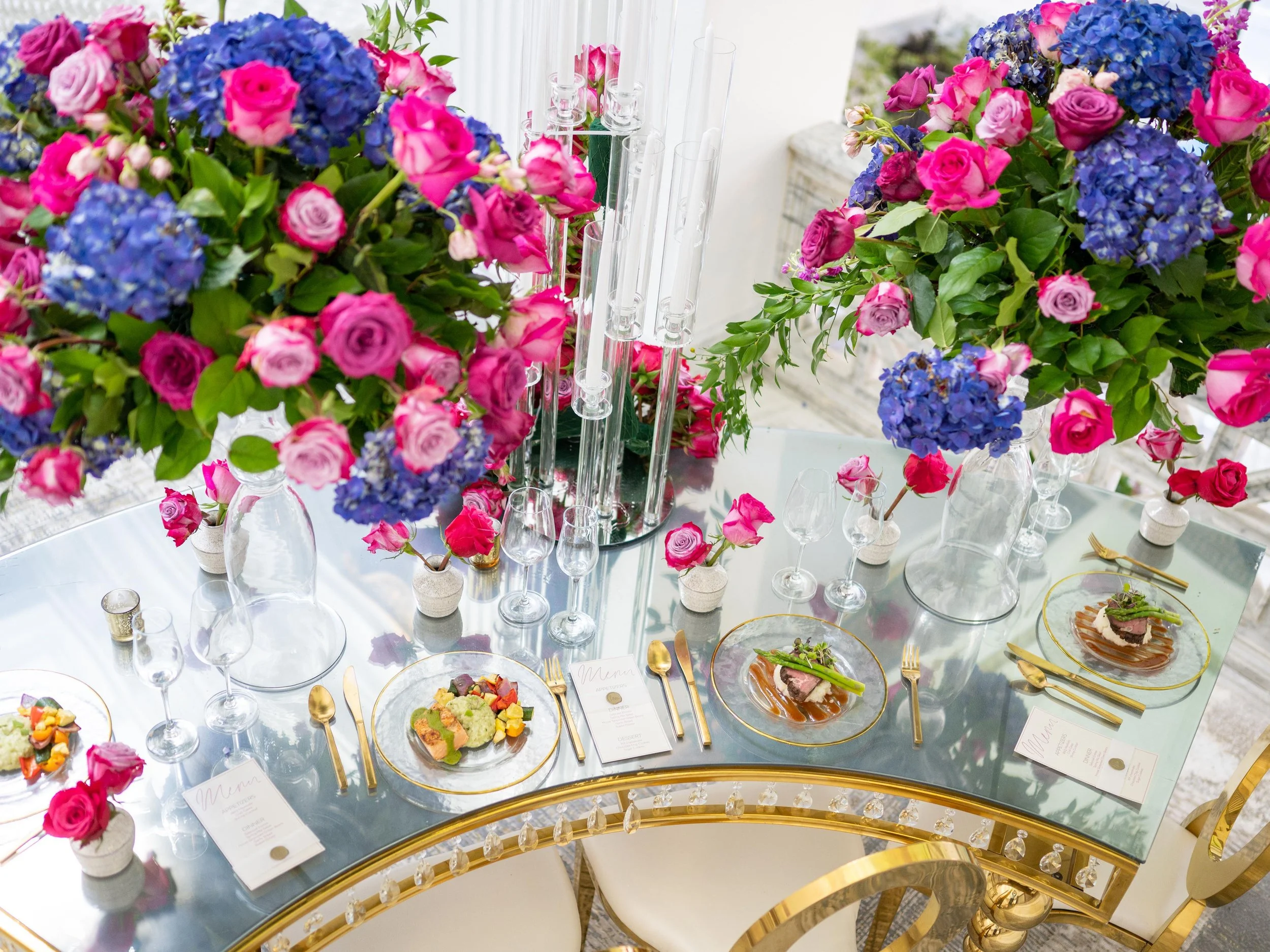 Elegant table setting with large floral centerpieces of pink roses and blue hydrangeas, gold-accented glassware, and place settings with gold utensils, white plates, and menus, on a glass-top table in a decorated dining area.
