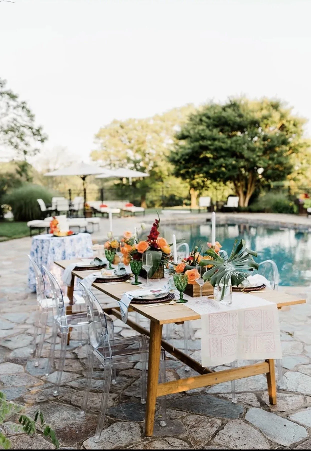 Outdoor dining table setup near a swimming pool with floral arrangements, transparent chairs, and a sunny backyard scene.